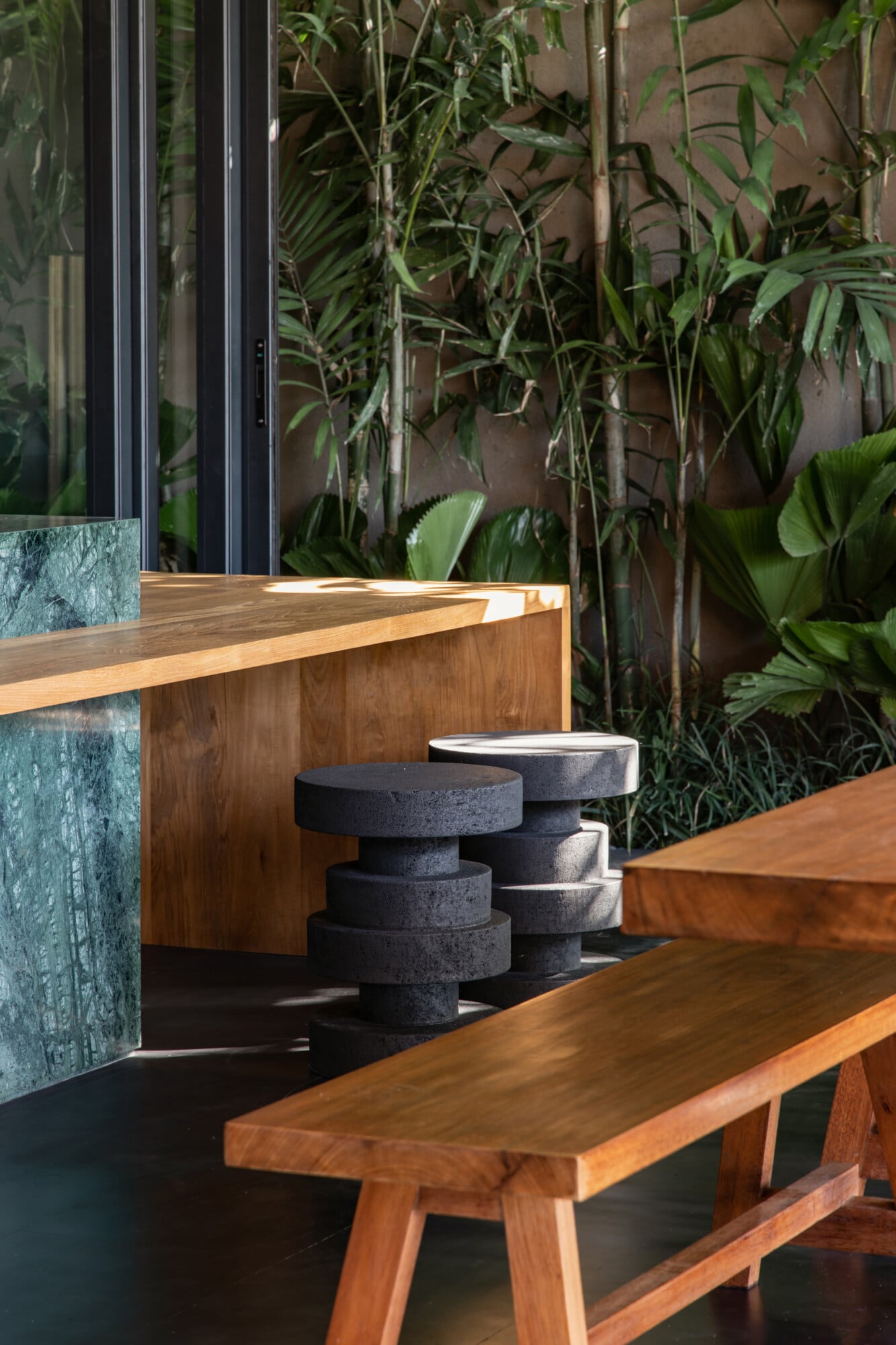 Tegel House, Asia-Pacific — Wooden bench and stacked charcoal stools beside angled timber counter, lush green foliage visible through glass doors, natural light casting shadows across dark flooring.