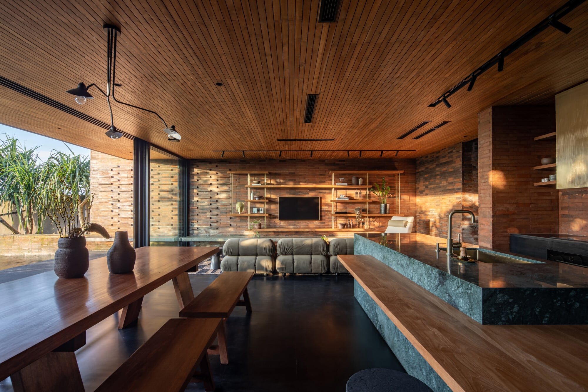 Tegel House, Asia-Pacific — Open-plan living with warm timber ceiling, brick walls, natural light, modern kitchen island, and structured shelving displaying curated objects.