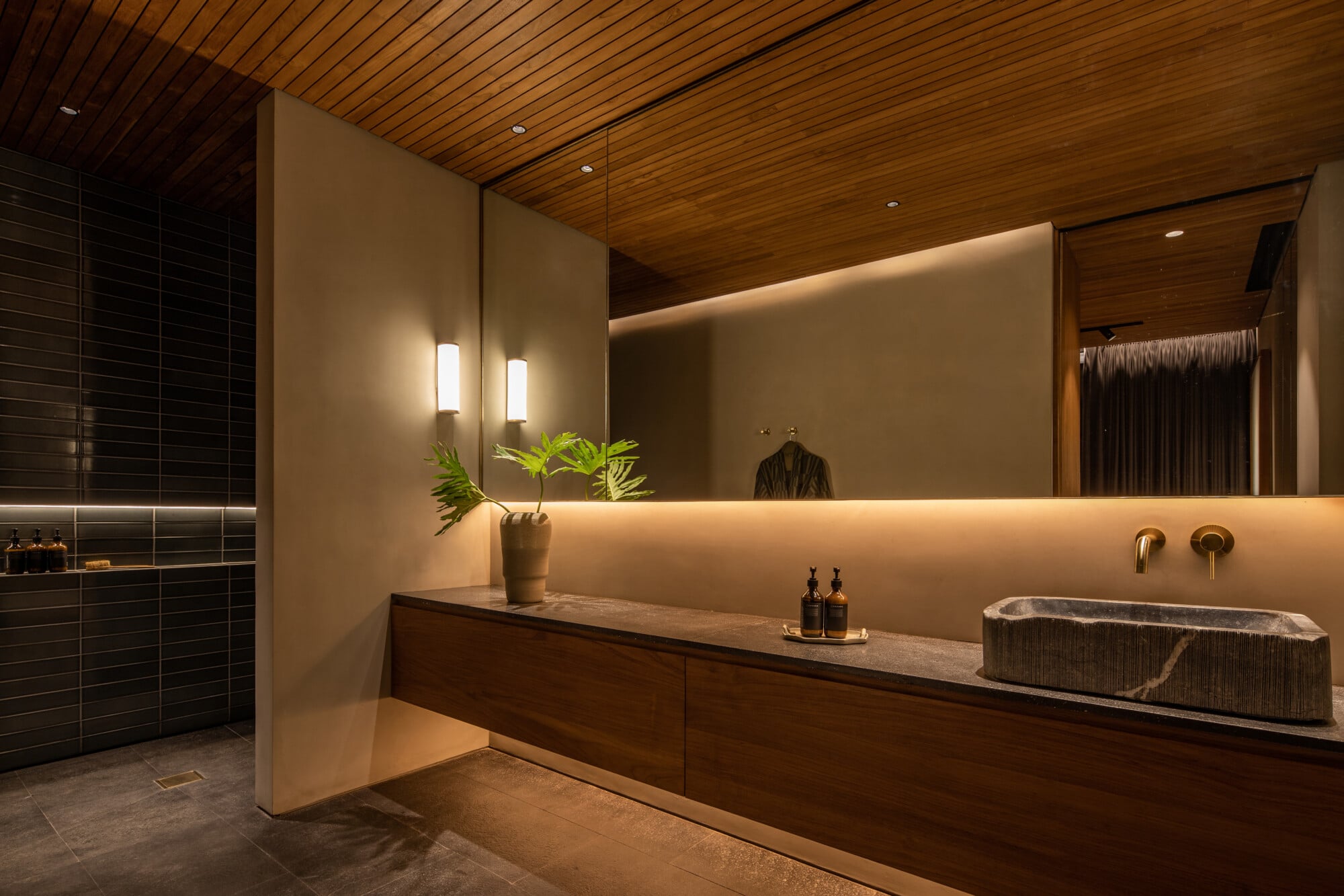 Tegel House, Asia-Pacific — Minimalist bathroom with walnut vanity, textured stone vessels, brass fixtures, backlit mirror, black tile shower, and wooden ceiling detail.