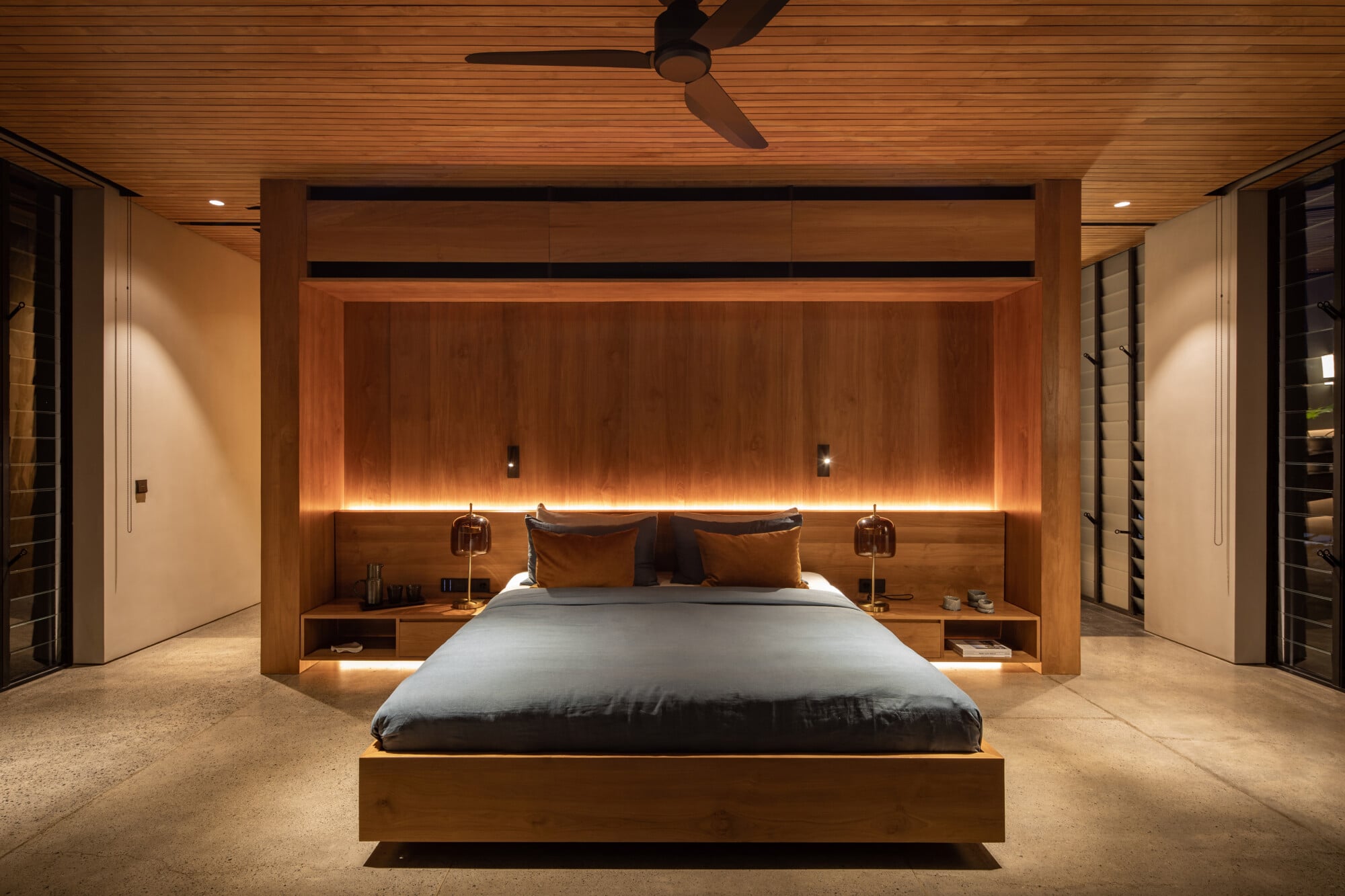 Tegel House, Asia-Pacific — Modern master bedroom with timber ceiling, floating bed, integrated wood headboard wall with ambient lighting, and minimalist pendant fixtures.