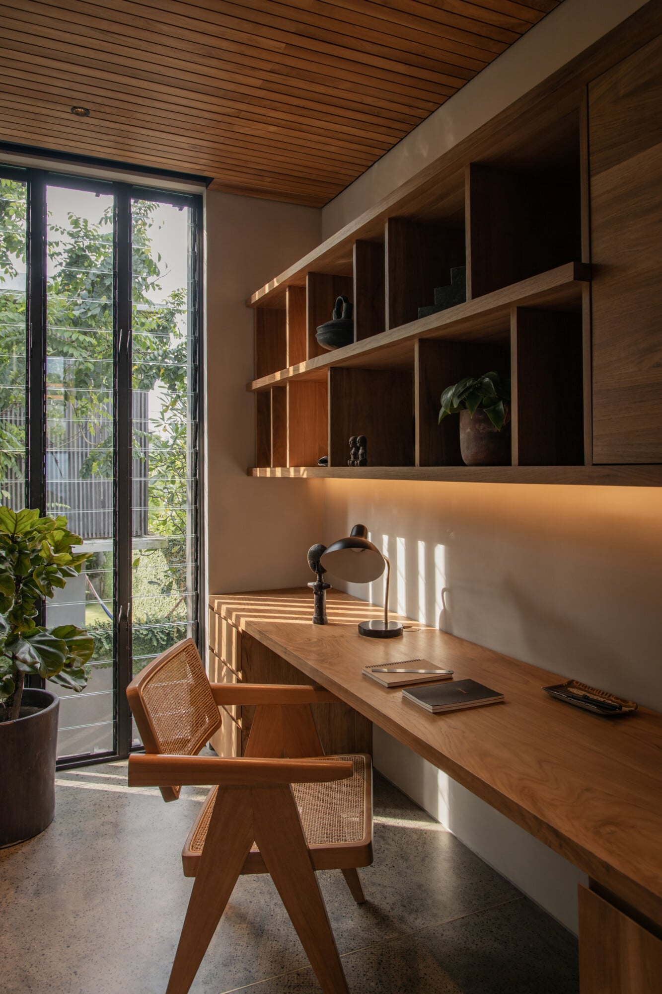 Tegel House, Asia-Pacific — Wooden desk workspace with built-in shelving, rattan chair, and floor-to-ceiling glass doors opening onto vegetation-filled balcony, natural light streaming through horizontal slats.