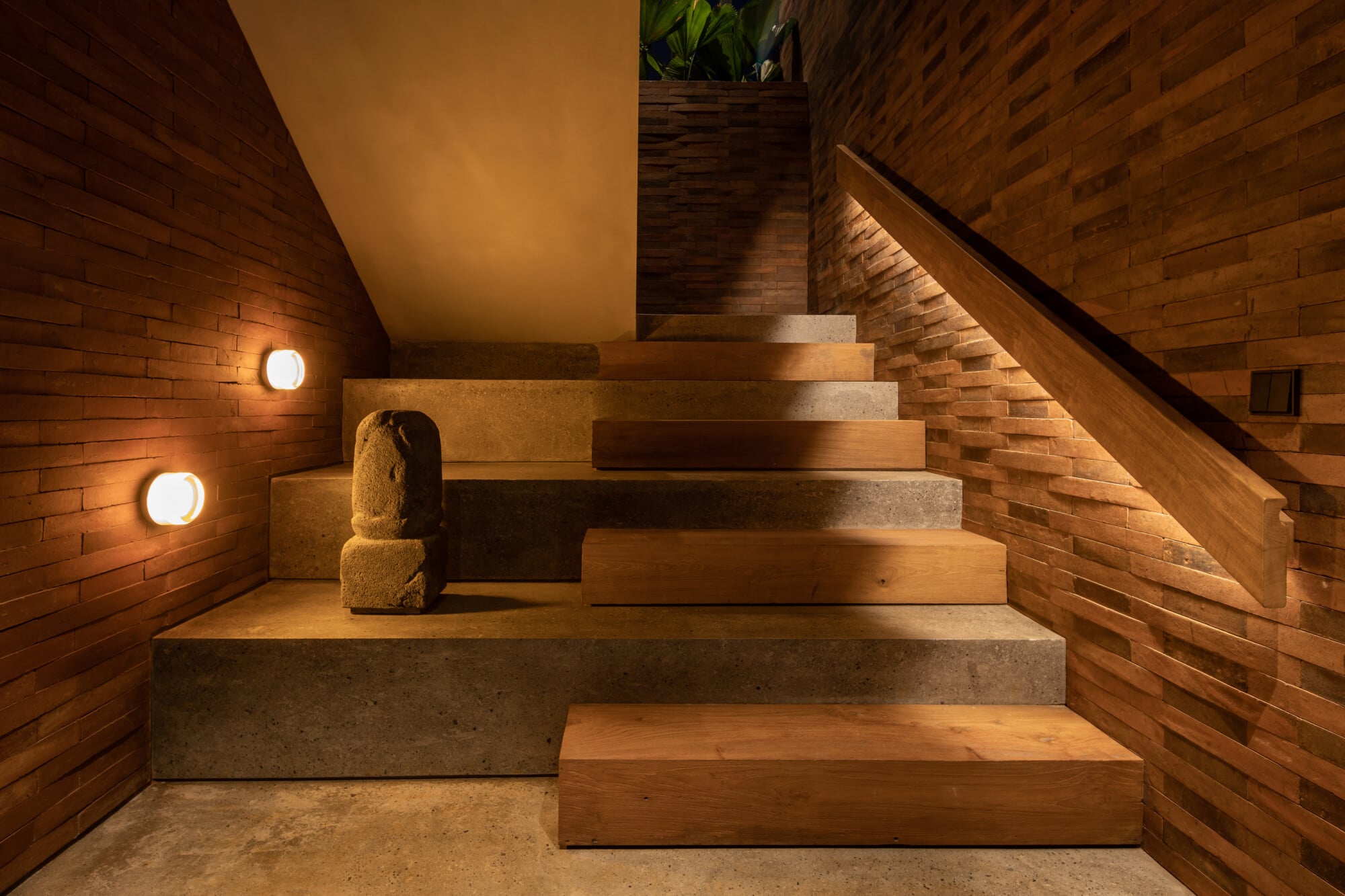 Tegel House, Asia-Pacific — Modern staircase with alternating wood and concrete steps, brick walls, recessed lighting, sculptural stone accent, and potted plants.