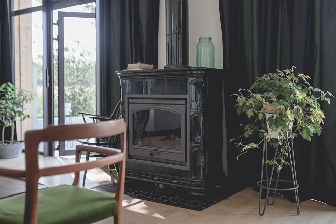 Le Barn, Europe — Modern black wood-burning stove beside floor-to-ceiling windows with dark curtains, potted plants, and mid-century wooden chair in minimalist interior.