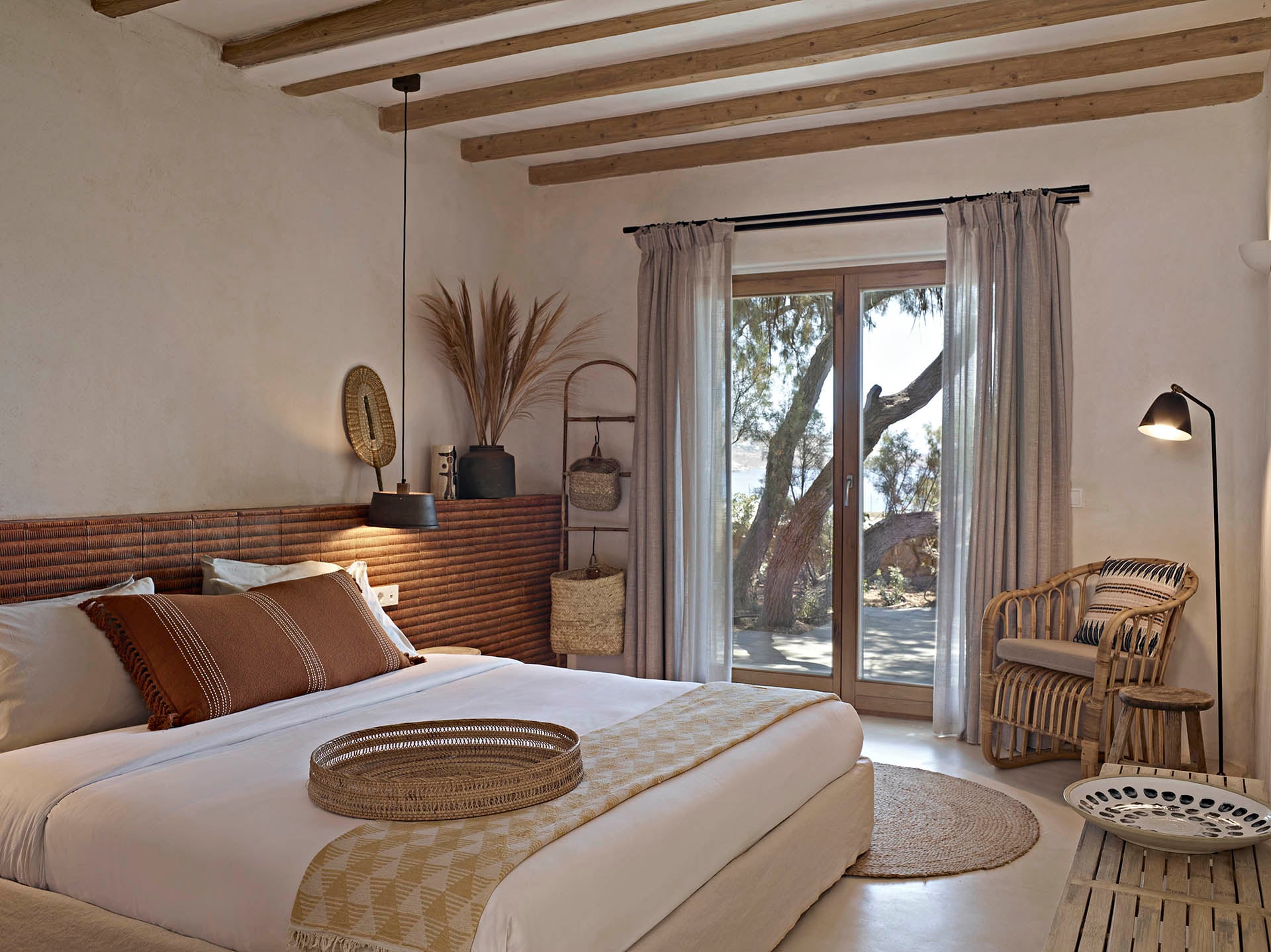 The Wild, Europe — Coastal bedroom with wooden beam ceiling, woven rattan chair, terracotta headboard, linen curtains framing glass doors overlooking olive tree landscape.