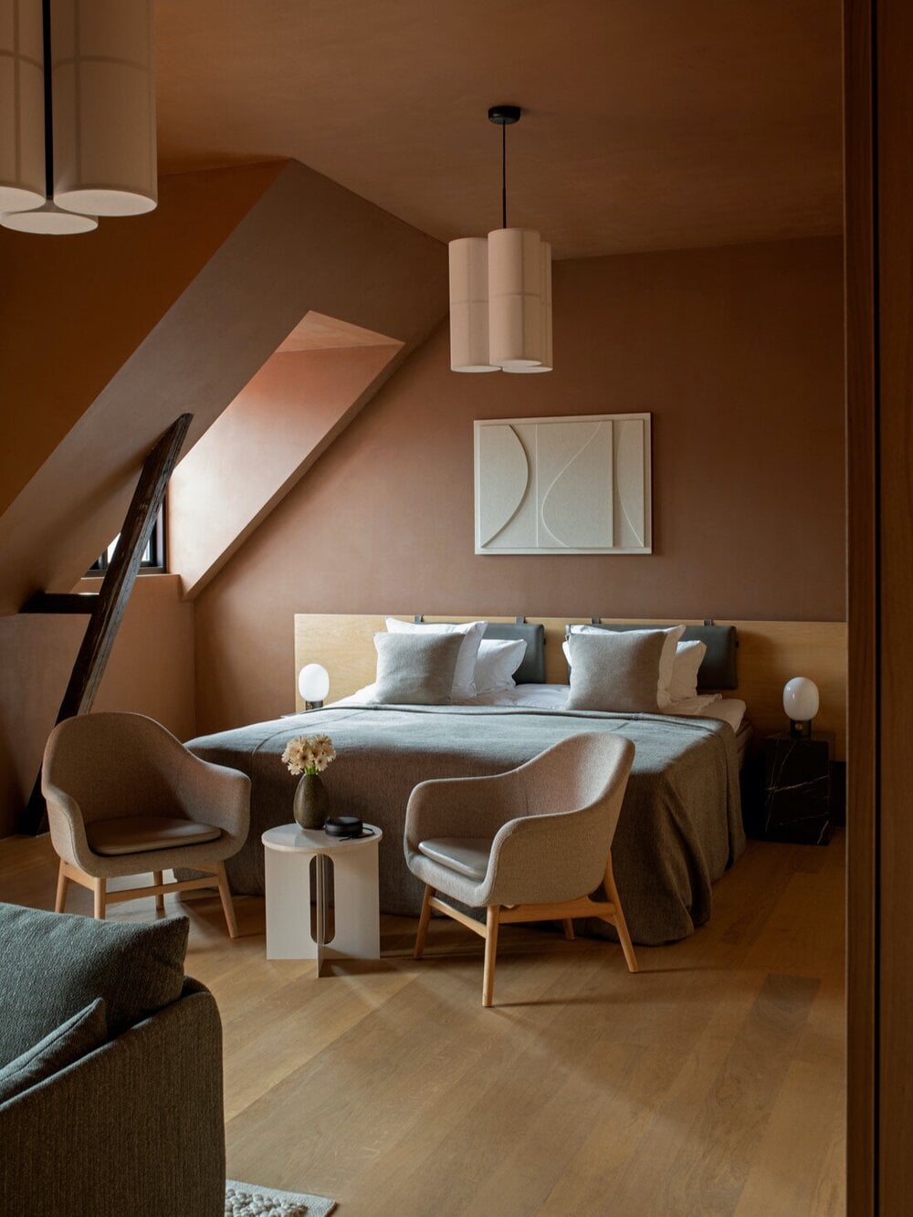 The Audo, Europe — Minimalist attic bedroom with terracotta walls, natural light, wooden furnishings, linen bedding, and mid-century modern chairs.
