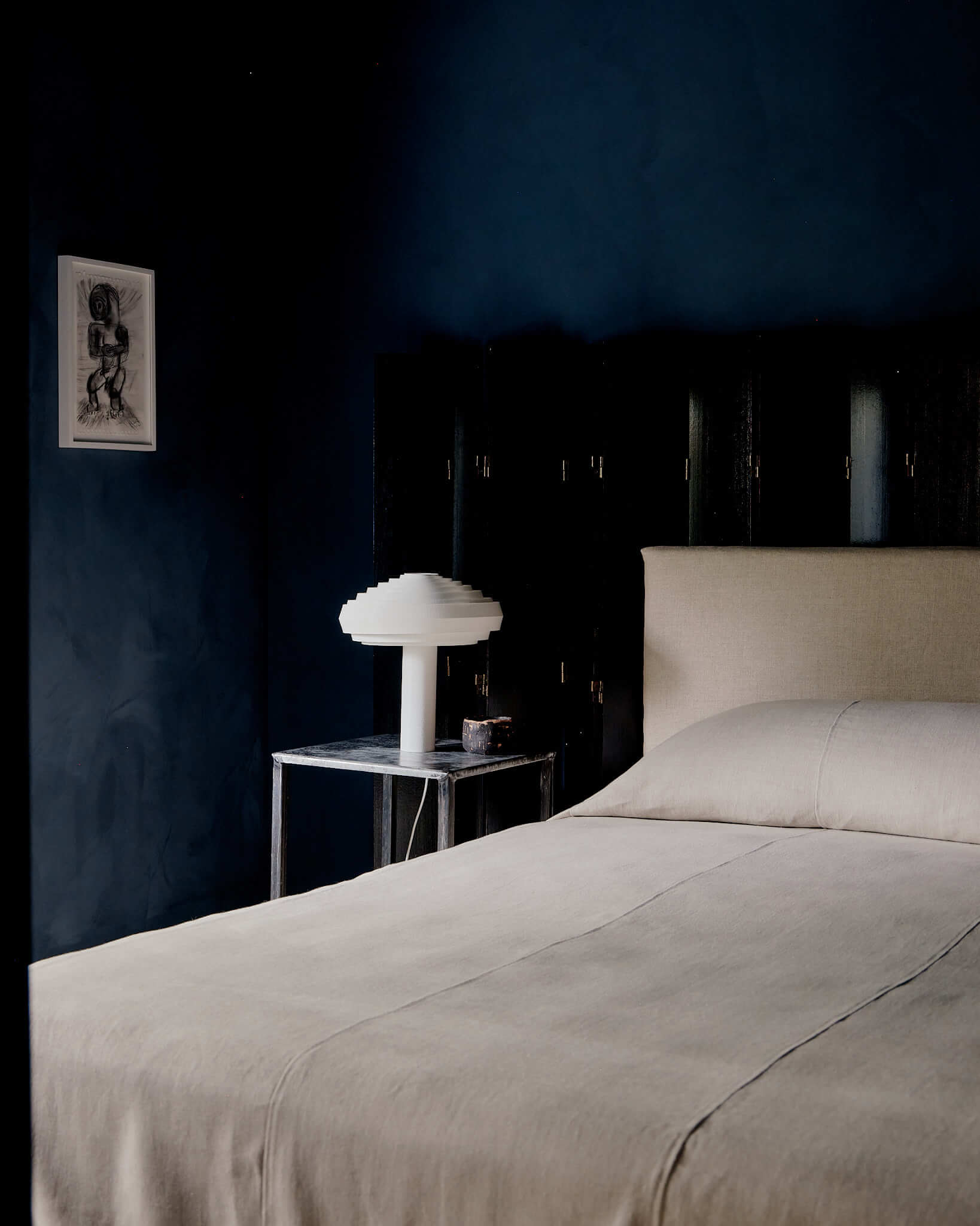 The James Bradley, North America — Minimalist bedroom with deep navy walls, cream bedding, sculptural white mushroom lamp, and framed artwork above dark wood nightstand.