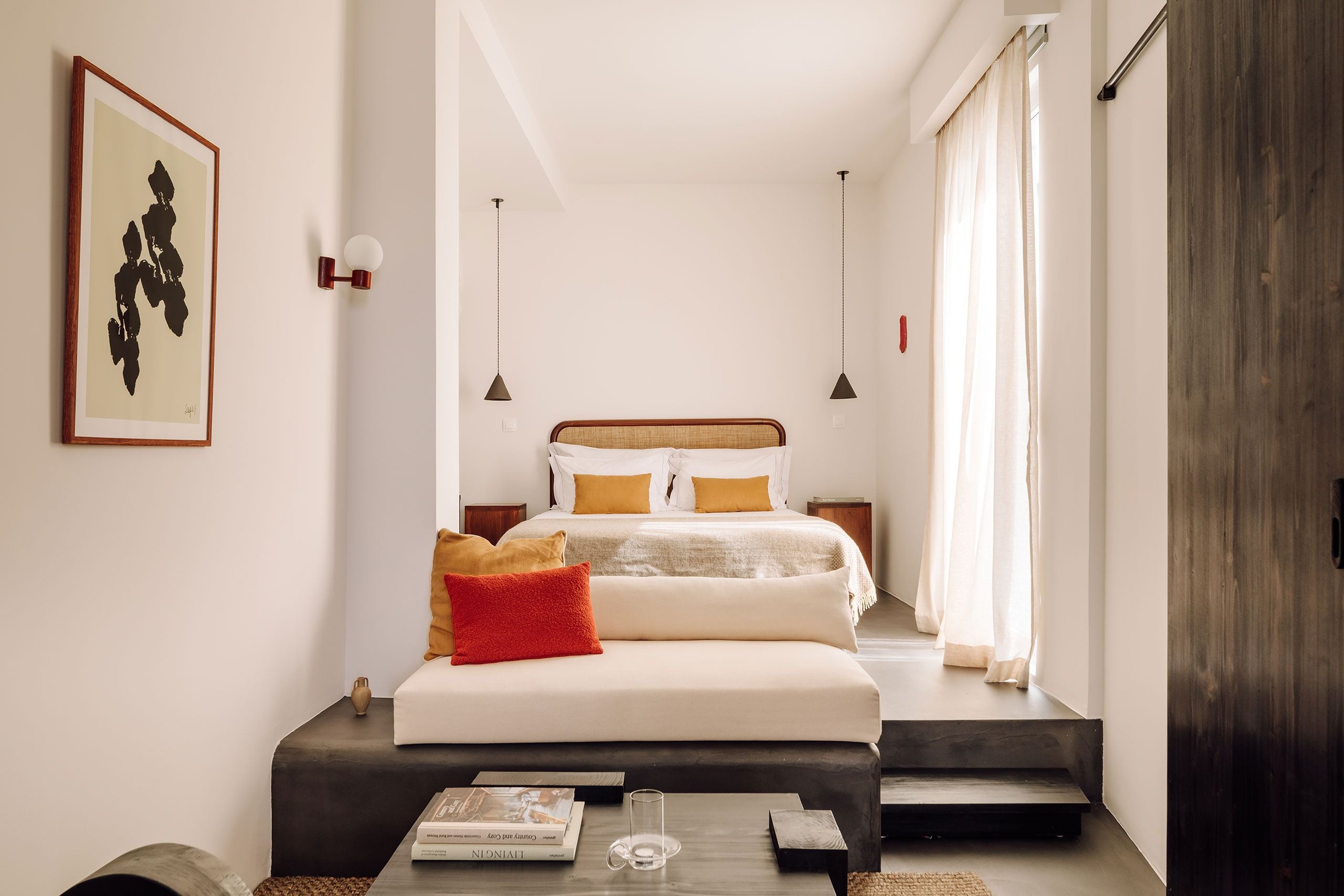White Hotel, Europe — Minimalist bedroom with rattan headboard, pendant lights, cream bedding, red accent pillows, dark wood bench, floor-to-ceiling windows, contemporary luxury hospitality design.
