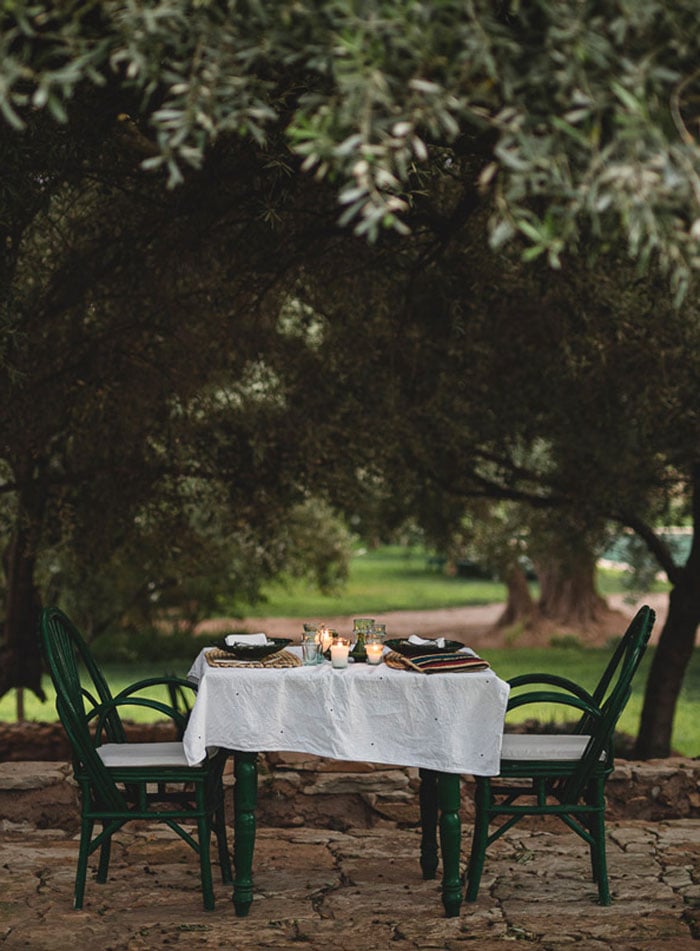 Berber Lodge, Africa — intimate outdoor dining beneath olive trees with white linen tablecloth, green bistro chairs, candlelight, and manicured grounds at dusk.