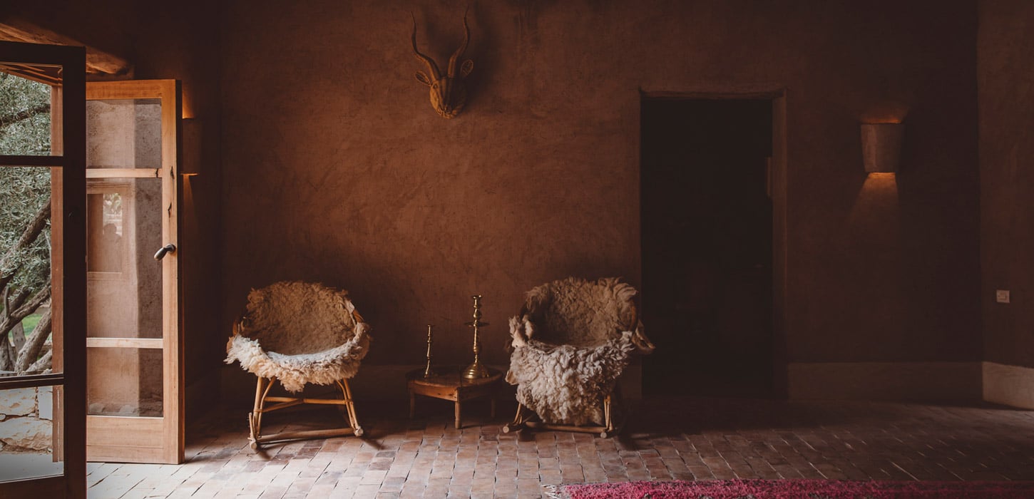 Berber Lodge, Africa — Ochre-walled room with sheepskin chairs, brass candleholder, terracotta floors, and warm natural light through open wooden door.