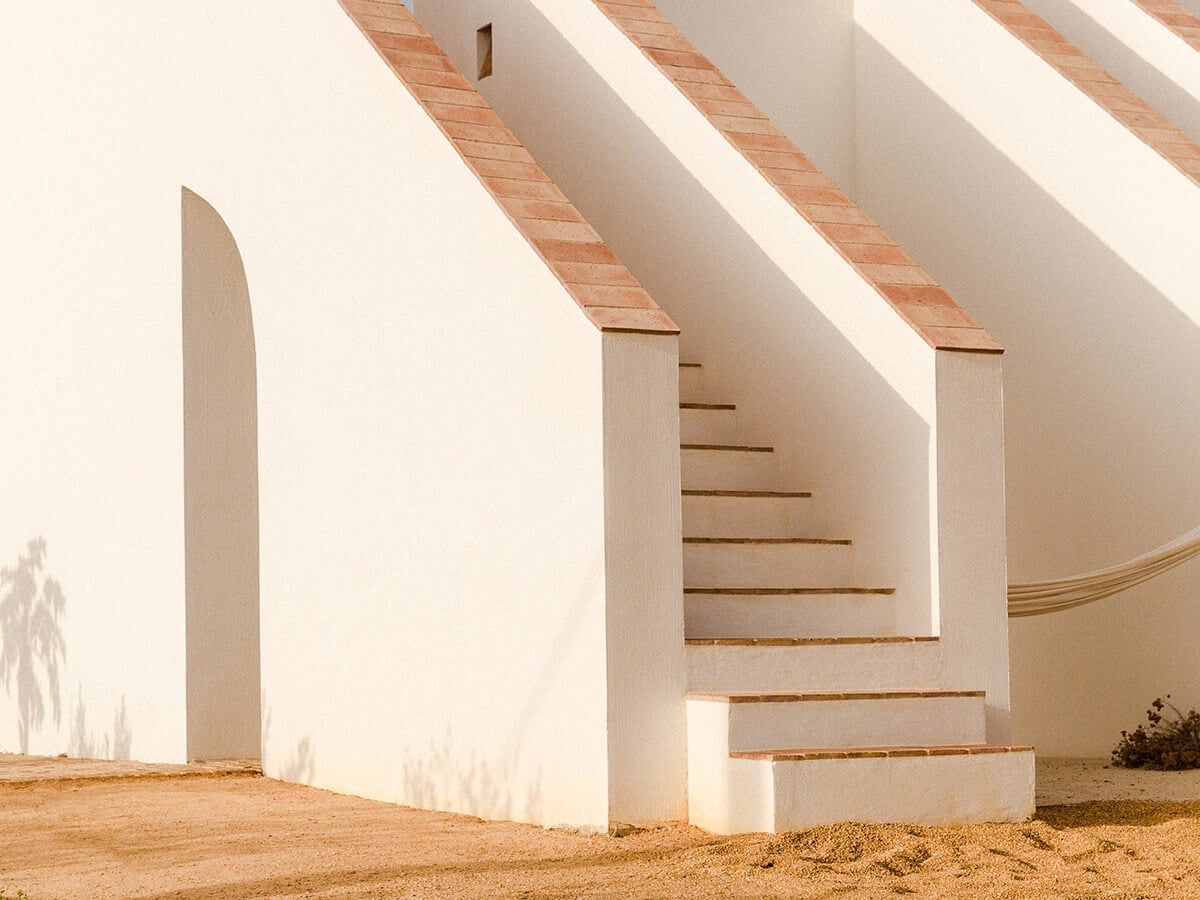 Casa Modesta, Europe — Minimalist white-walled staircase with timber treads and brick detailing, casting sharp shadows in desert sunlight.
