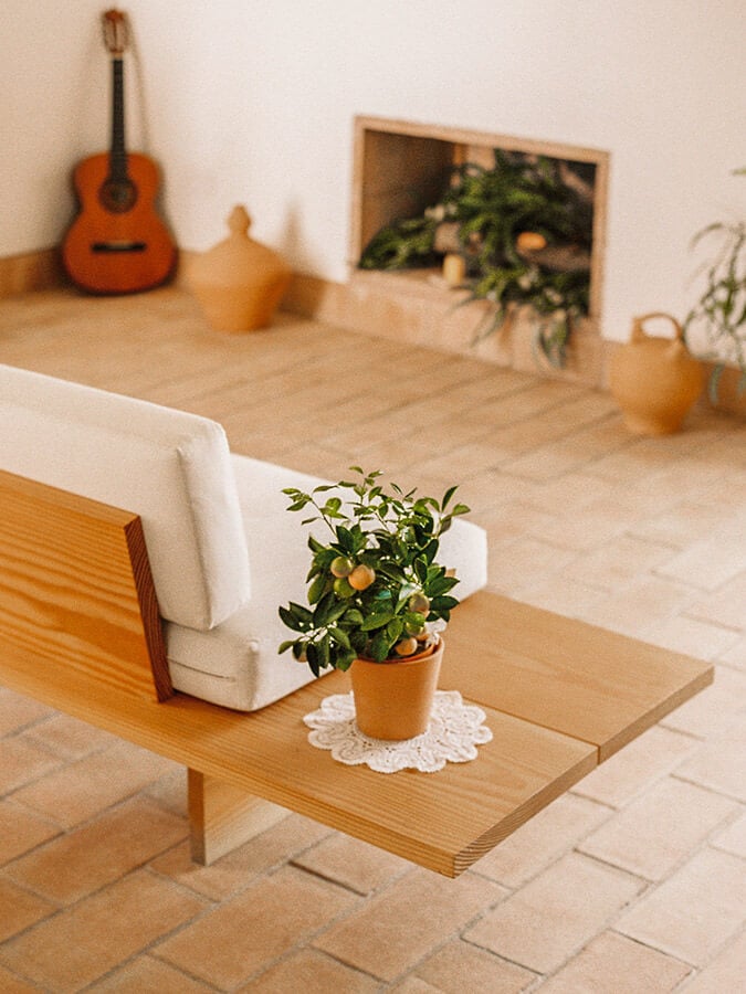Casa Modesta, Europe — Minimalist wooden table with potted citrus plant, white cushioned chair, guitar, and ceramic vessels in warm-toned interior space.