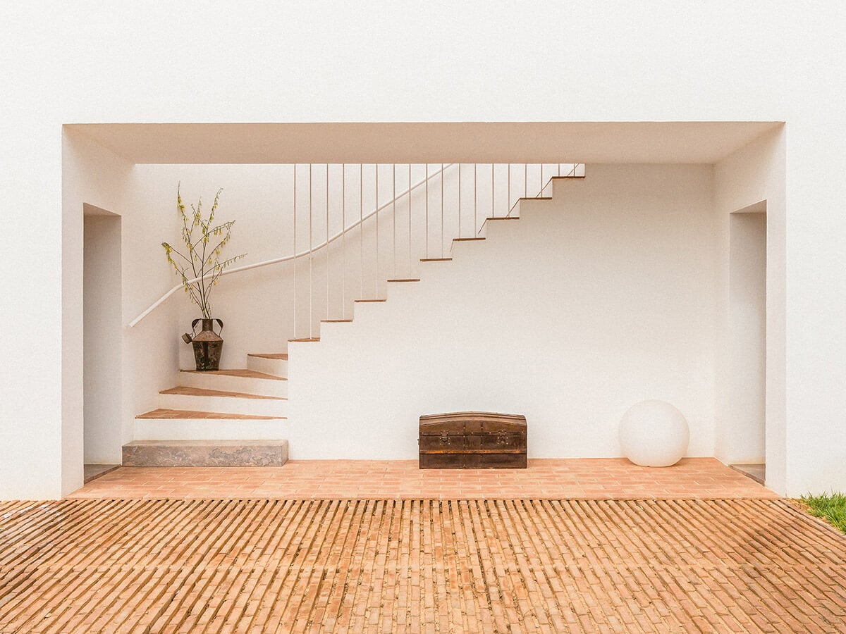 Casa Modesta, Europe — Minimalist interior with floating wooden staircase, metal railings, terracotta brick floor, white walls, and curated vintage objects.