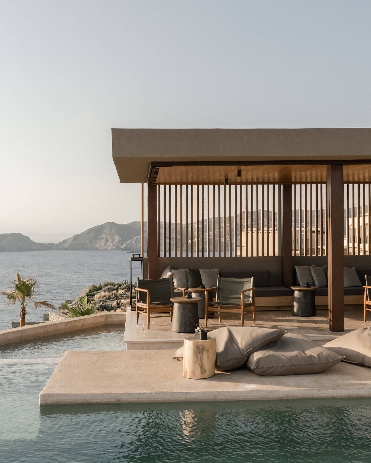 ACRO Suites, Europe — Waterfront pavilion with wooden slatted screens, stone terraces, teak lounge chairs, and sea views at dusk.