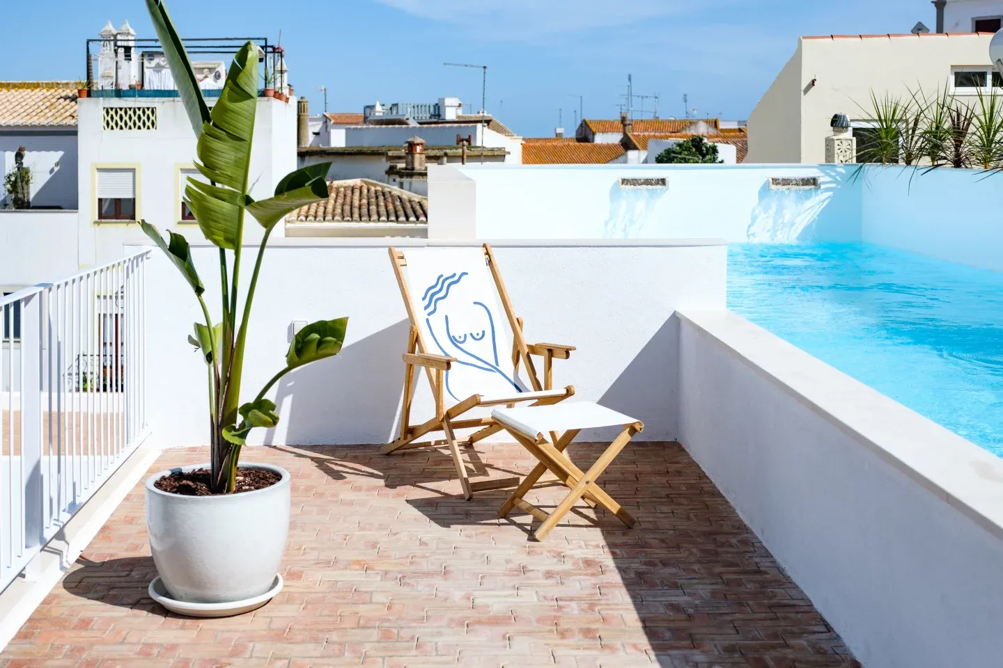 Casa Mãe, Europe — Rooftop terrace with wooden lounger, potted banana plant, and mural overlooking whitewashed Mediterranean architecture under clear blue sky.