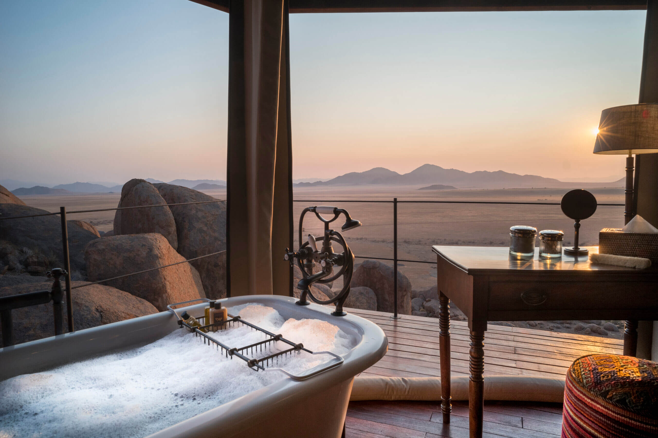 Zannier Sonop, Africa — Soaking tub with sunset lake view, granite boulders, wooden deck, bicycle art installation, mountains on horizon at dusk.