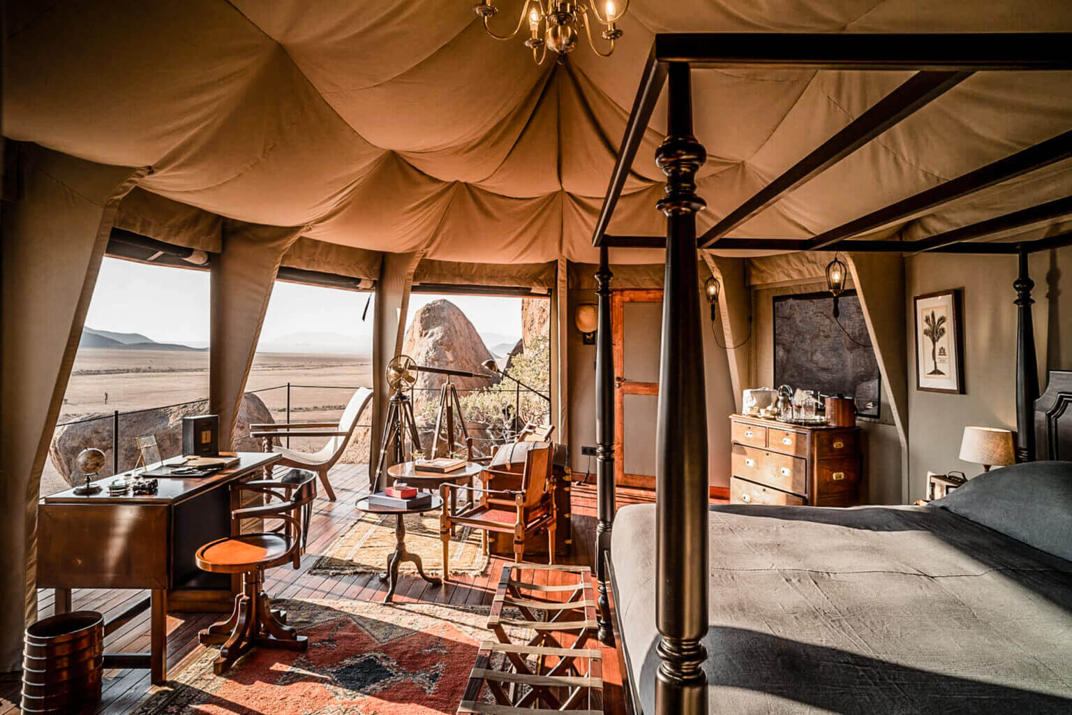 Zannier Sonop, Africa — Canvas-draped luxury tent interior with wooden furnishings, brass four-poster bed, and expansive desert landscape views through floor-to-ceiling windows.