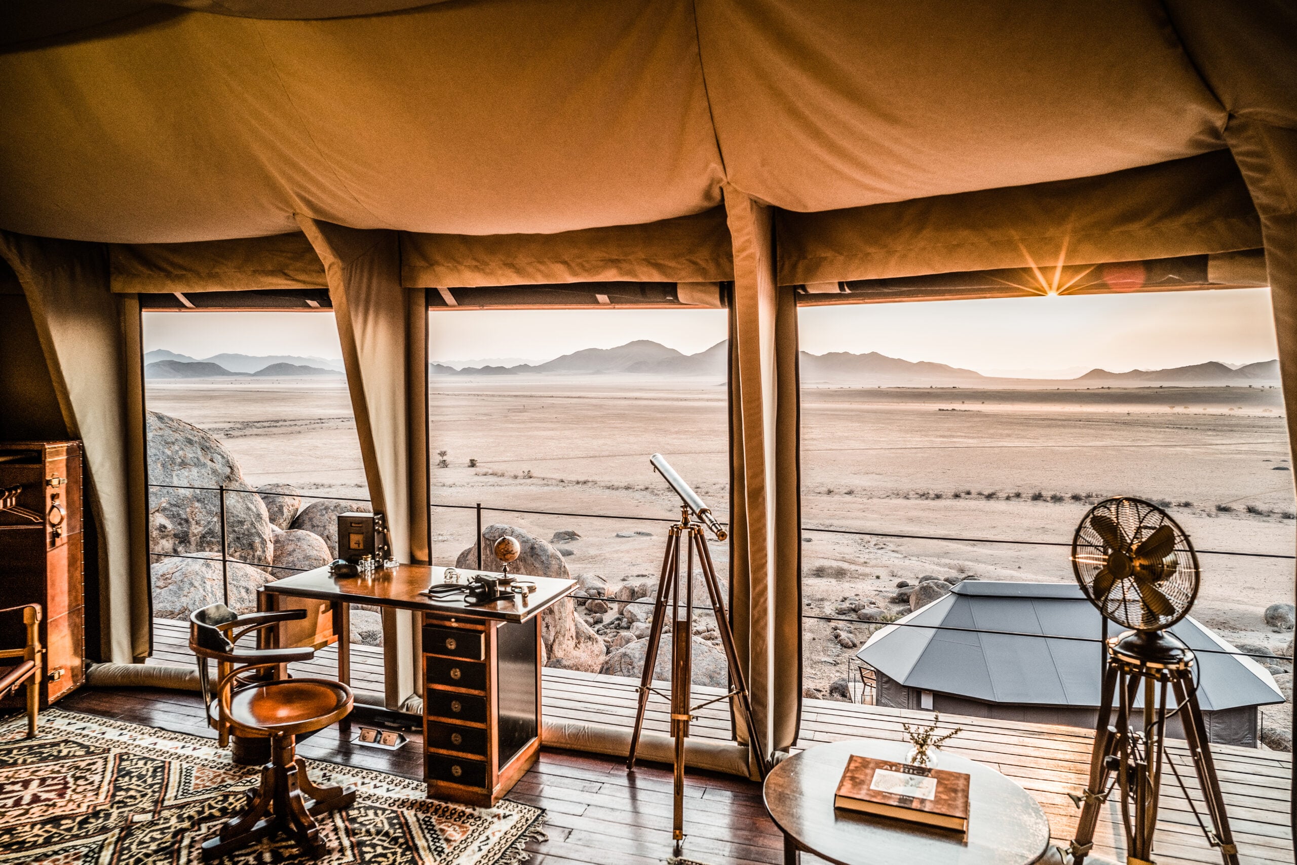 Zannier Sonop, Africa — Luxury safari tent interior with wooden desk, telescope, and expansive desert landscape views of mountains and plains.