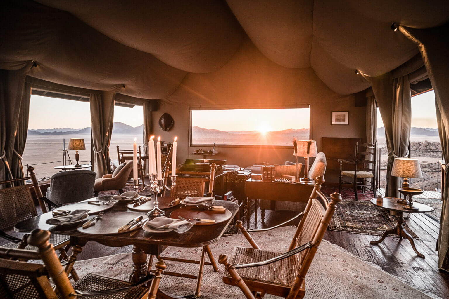 Cocktail & Cigar Lounge — Luxury safari lodge lounge with canvas ceiling, wooden furnishings, sunset views over African landscape, candlelit dining setup, and coastal vista.