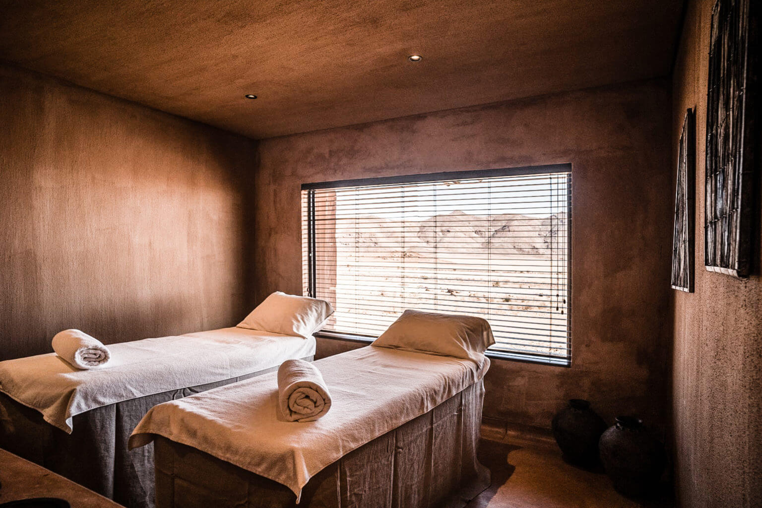 Sonop Spa, Africa — Minimalist spa treatment room with terracotta walls, two massage tables with rolled towels, horizontal wooden blinds filtering warm light, earthy material palette.