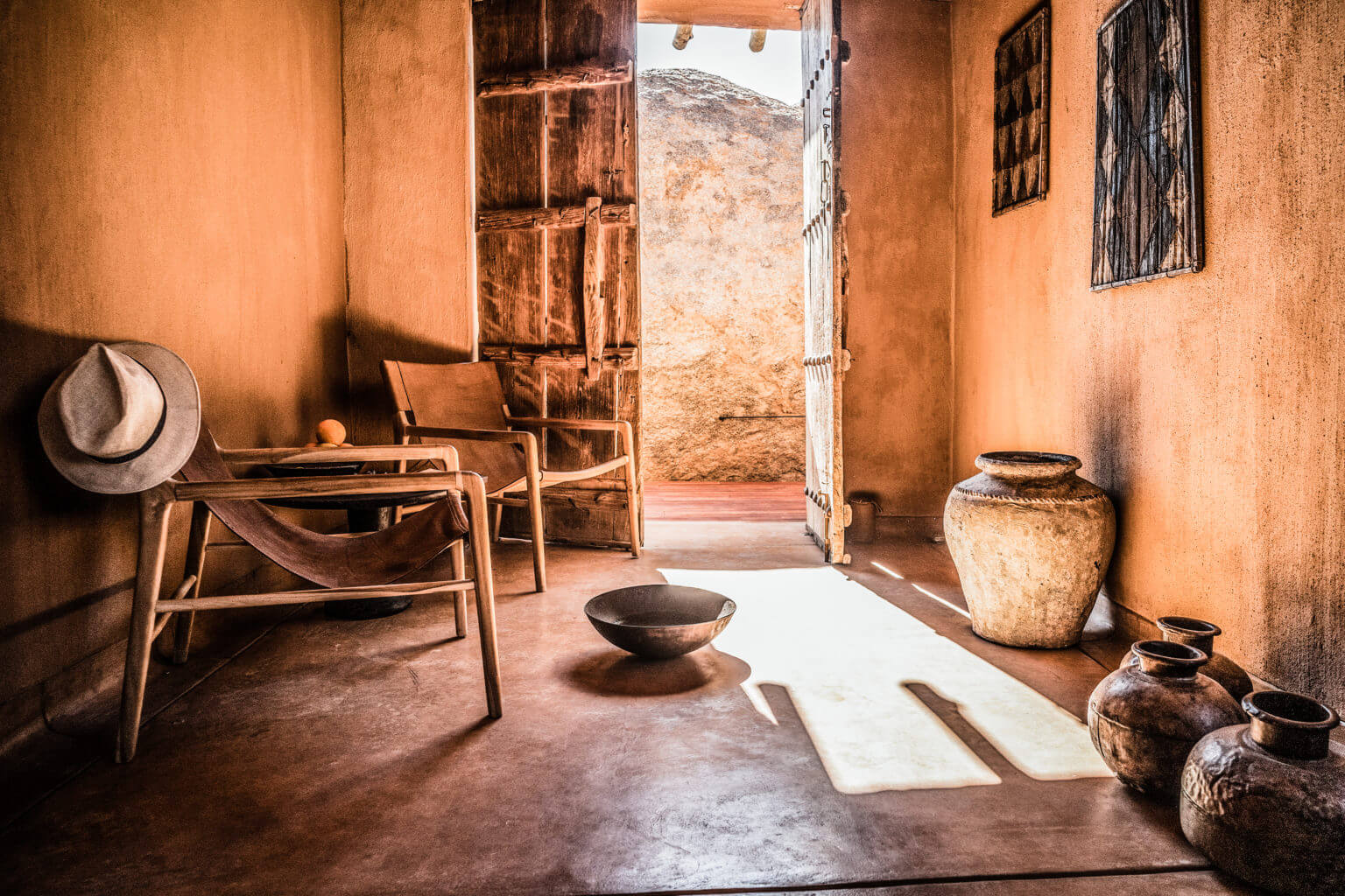 Zannier Sonop, Africa — Terracotta interior with wooden chairs, ceramic vessels, open door framing desert landscape, natural light casting shadows on earthen floor.