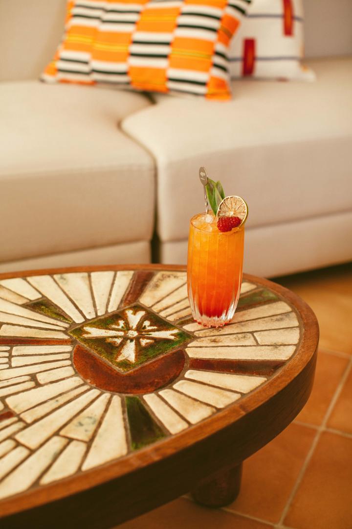 Hôtel Lou Pinet, Europe — Orange cocktail with lime and cherry on geometric wood side table, striped pillows and cream sofa in background.