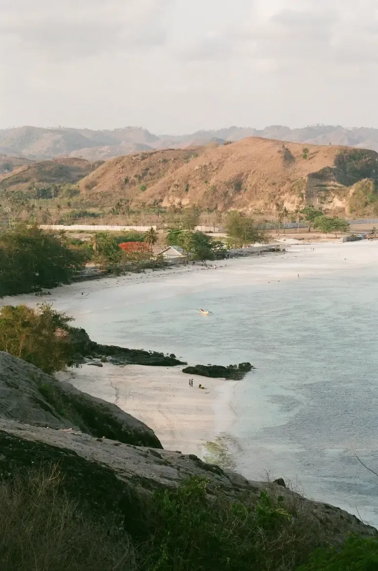 Somewhere Lombok, Indonesia — Turquoise bay with white sand beach, weathered volcanic hillside, tropical vegetation, and overcast sky at golden hour.