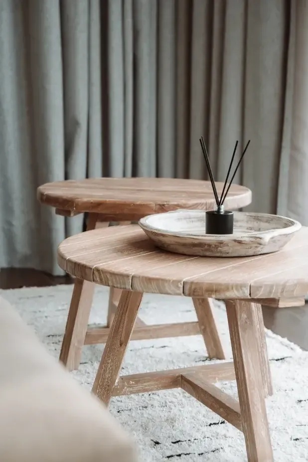 Somewhere Lombok, Asia — Light wood round tables with natural grain detail, ceramic dish with black reed diffuser, minimalist styling against linen curtains.