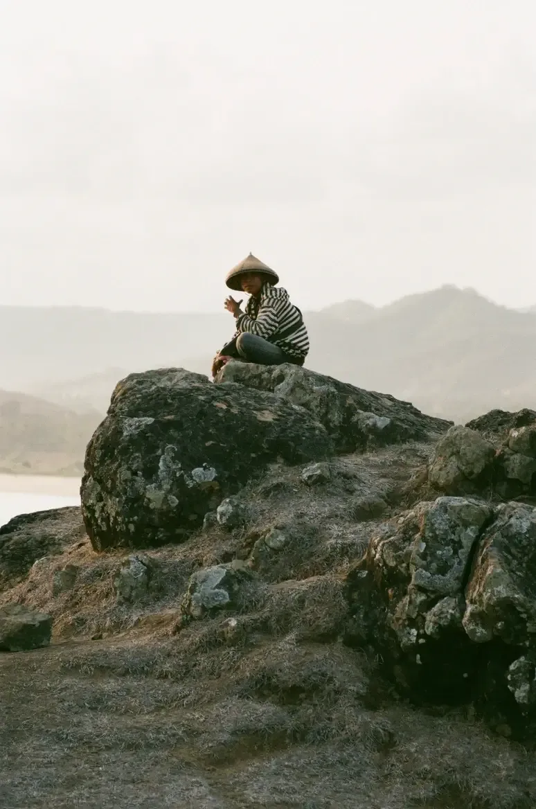 Somewhere Lombok, Asia — Person in conical hat seated on lichen-covered boulder overlooking misty mountains and water, striped shirt, contemplative mood.