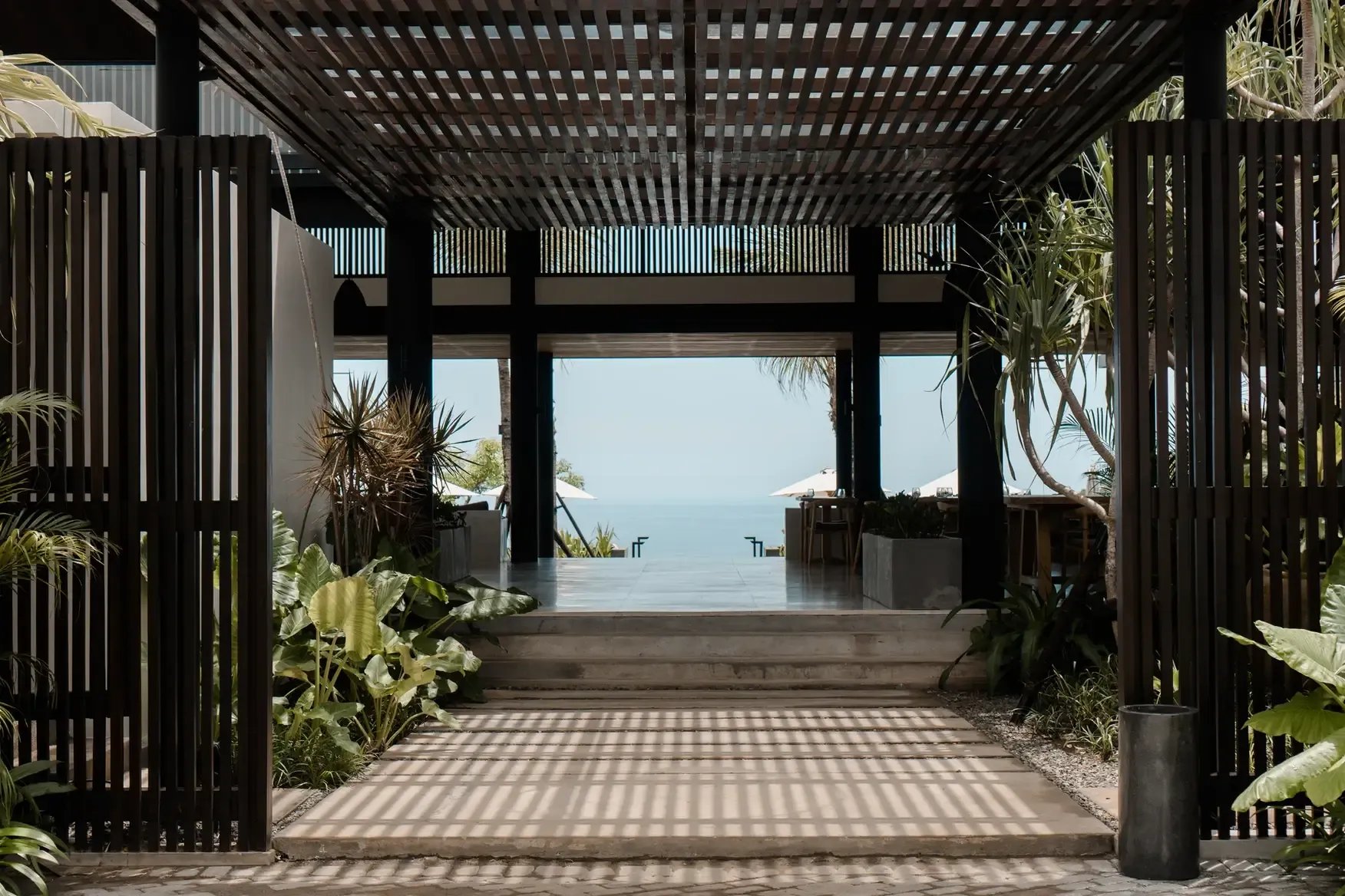 Somewhere Lombok, Asia — Dark timber pergola frames ocean view through slatted screens, tropical plants, and wooden deck with dappled light and sea horizon.