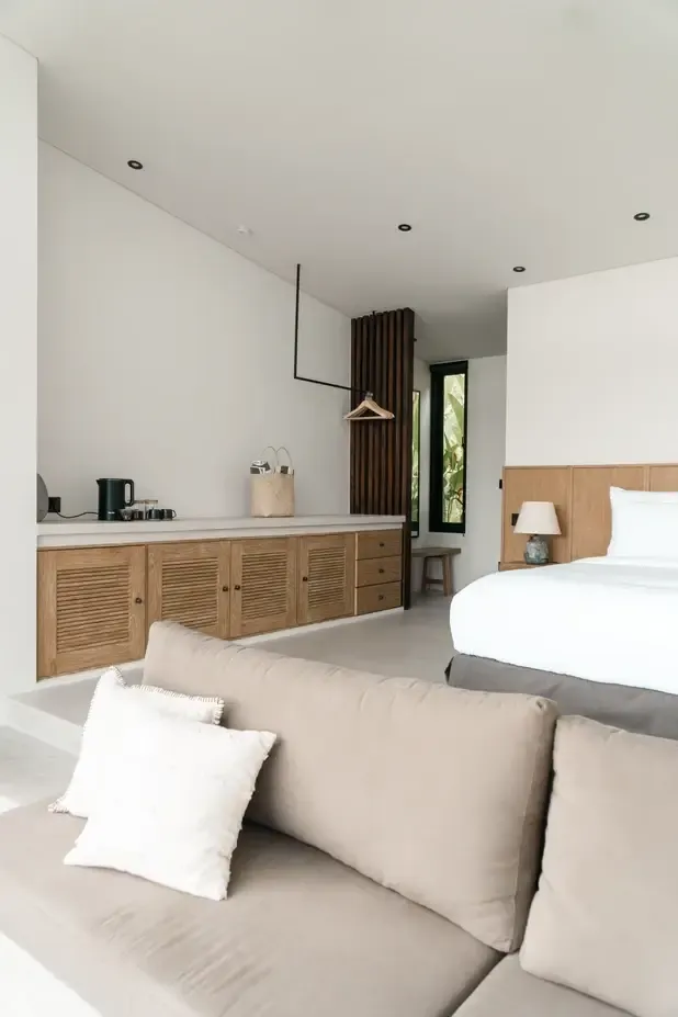 Somewhere Lombok, Asia — Modern villa bedroom with teak louvered cabinetry, white linens, brown curtains, concrete flooring, and minimalist coastal design aesthetic.