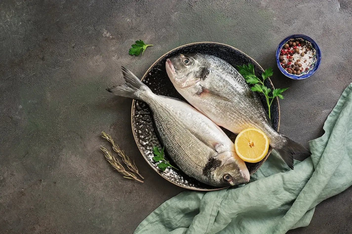 Somewhere Lombok, Asia — Fresh whole fish arranged on dark ceramic plate with lemon, herbs, and peppercorns on textured gray surface, styled for luxury hospitality dining.