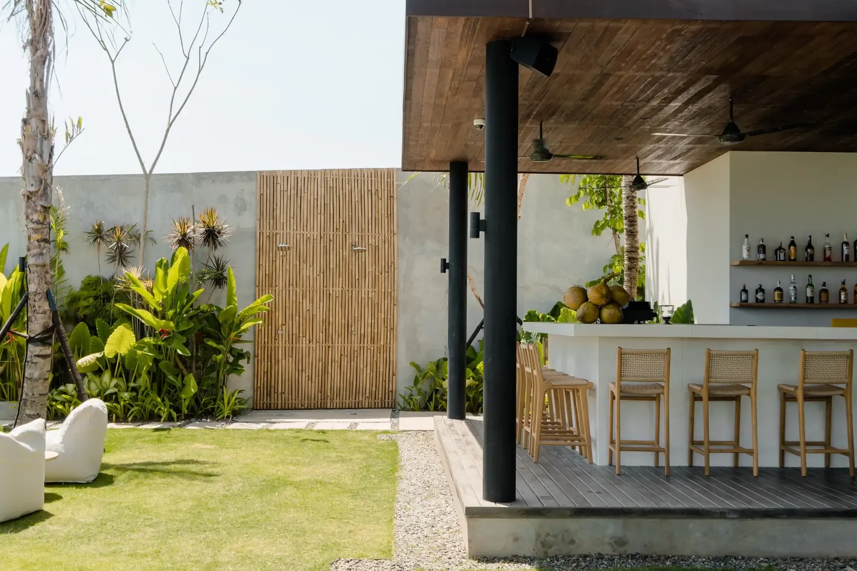 Somewhere Lombok, Bali, Indonesia — Modern tropical bar and garden courtyard with wooden pergola, palm trees, concrete walls, rattan seating, and lush landscaping.