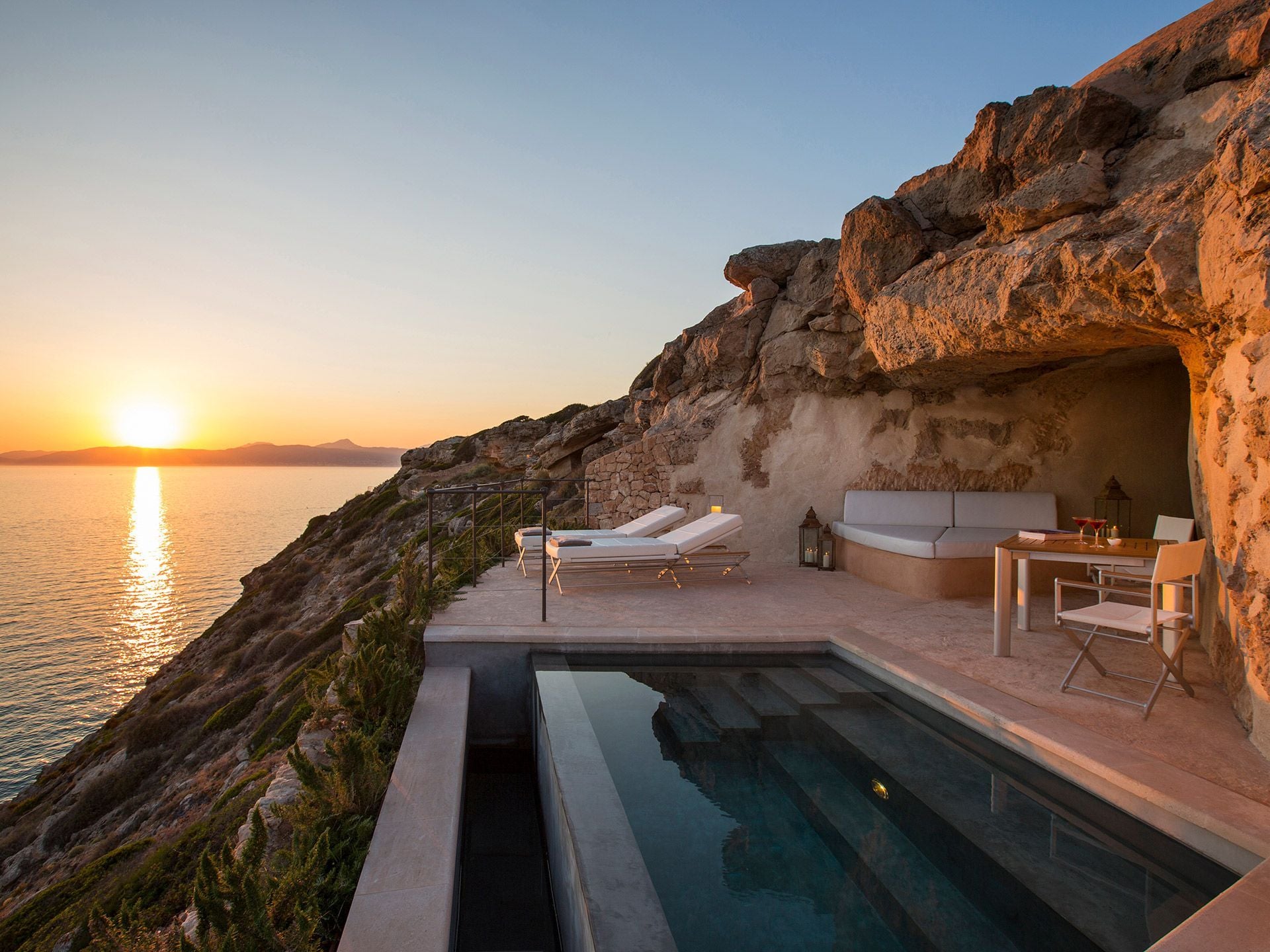 Cap Rocat, Balearic Islands — Clifftop pool terrace at sunset with limestone cave architecture, modernist loungers, and Mediterranean sea vista.