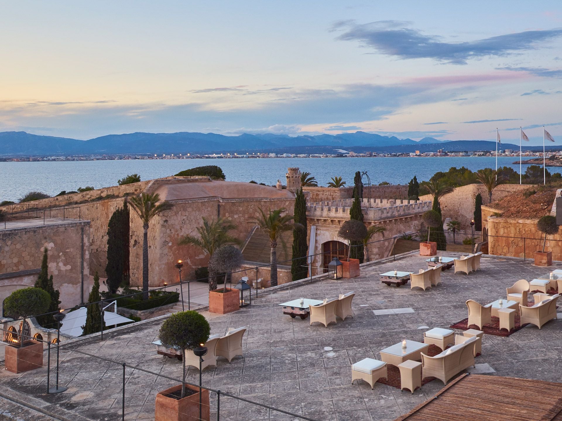 Cap Rocat, Balearic Islands, Spain — Stone fortress terrace with cream loungers overlooking Mediterranean bay at dusk, mountains and coastal town beyond.