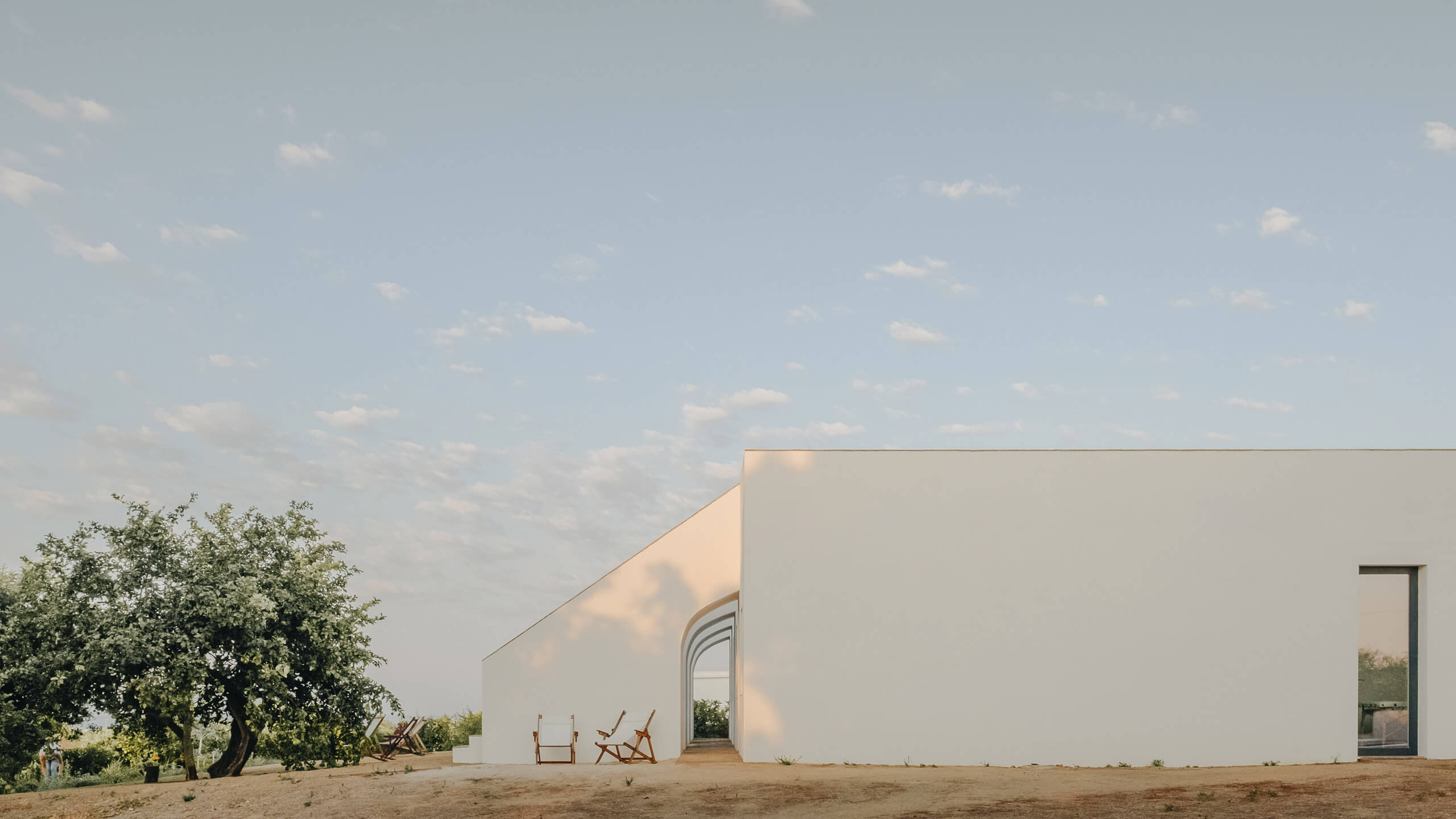 Casa Modesta, Europe — Minimalist white-walled architecture with curved entryway, wooden lounge chairs, mature tree, and sandy courtyard under clear sky.