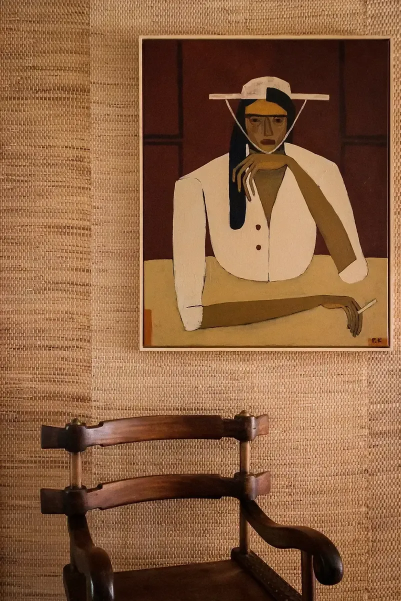 Sterrekopje, Africa — Modernist portrait painting of figure in cream blazer and wide-brimmed hat against terracotta wall, displayed above wooden chair.