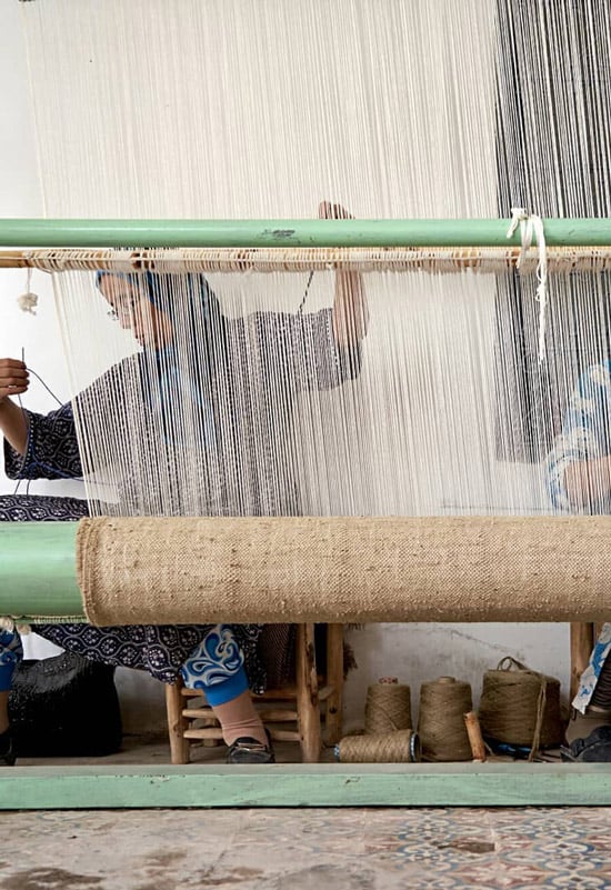 Wearing workshop near hotel, Africa — weaver at traditional loom with natural fiber threads and green wooden frame, artisanal textile craftsmanship.