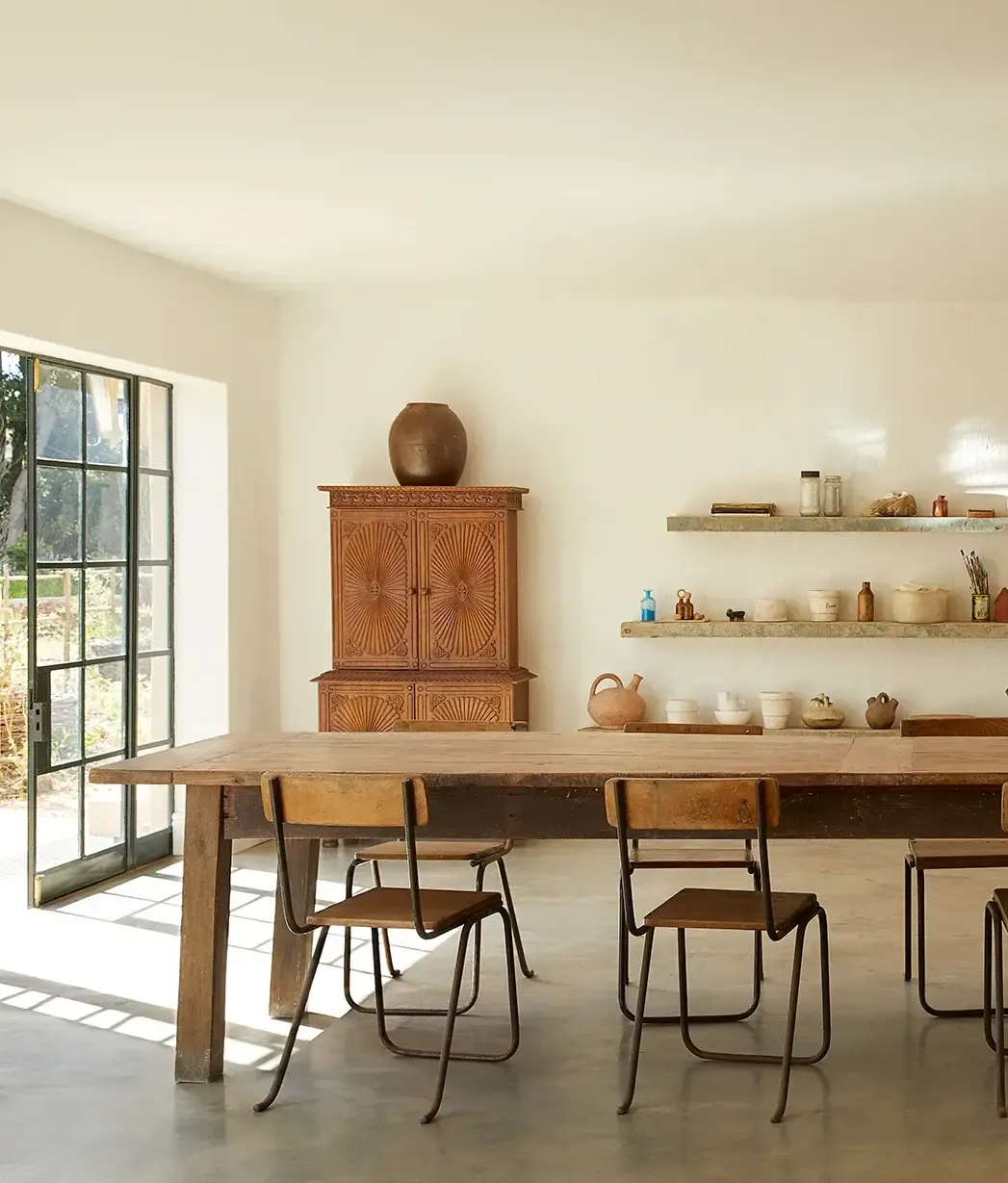 The Atelier, Africa — Minimalist dining space with wooden table, metal chairs, carved cabinet, floating shelves, and gridded glass doors opening to natural light.