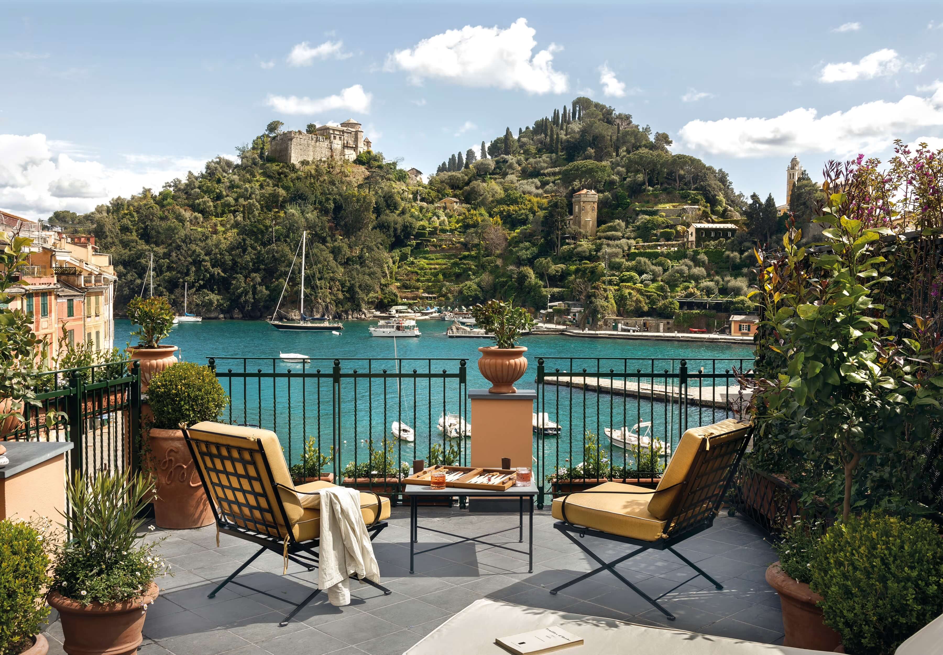 Splendido Mare, Europe — Waterfront terrace with ochre lounge chairs overlooking turquoise bay, hillside villa, cypress trees, and moored sailboats under blue sky.