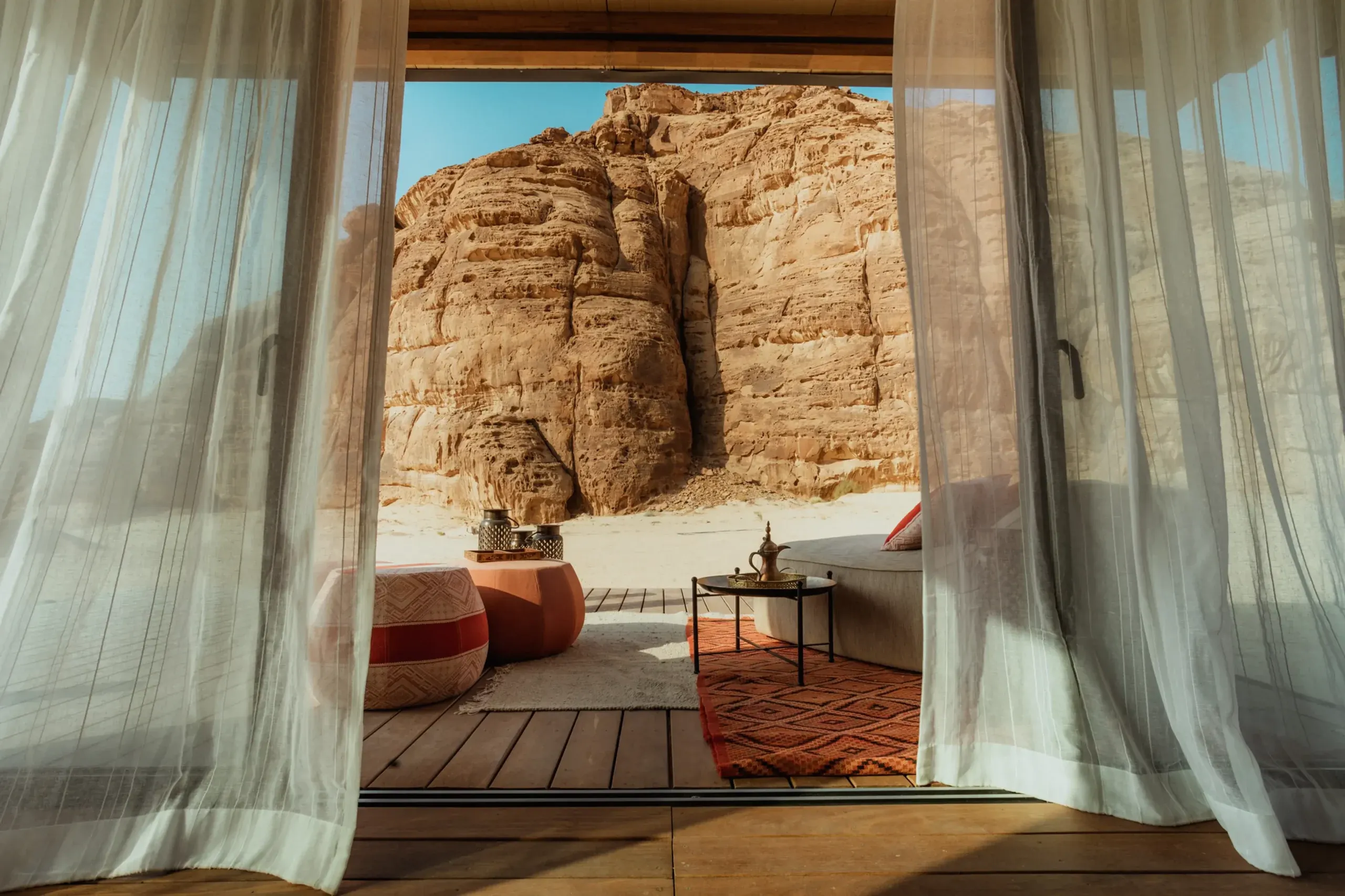 Habitas AlUla, Saudi Arabia — Open terrace with sheer curtains framing sandstone cliffs, geometric rugs, and modern furnishings against desert landscape.