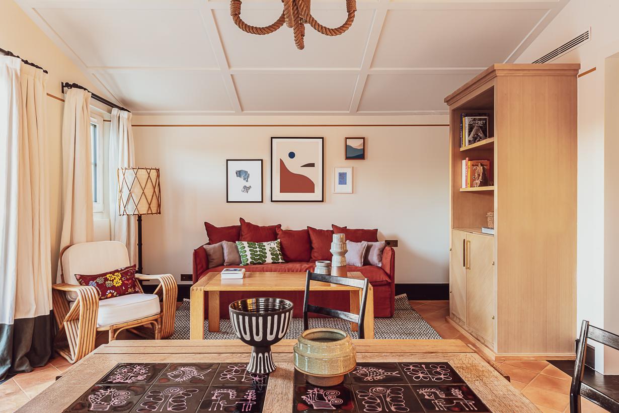 Hôtel Lou Pinet, Europe — Warm living room with red sofa, striped armchair, wooden shelving, coffered ceiling, and geometric patterned rug in neutral tones.