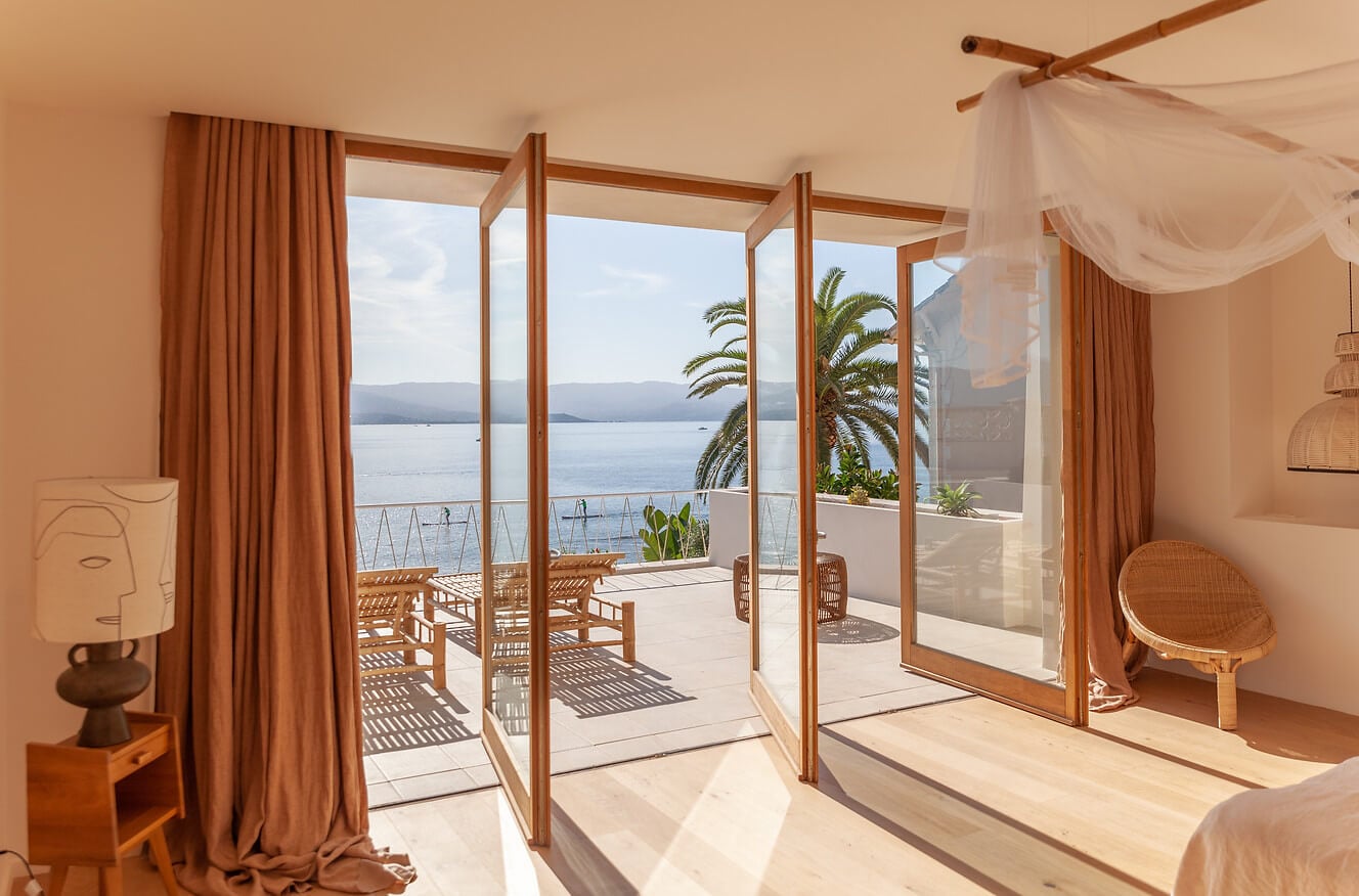 Casa Santa Teresa, Europe — Waterfront bedroom with wooden-framed glass doors opening to terrace, ochre linen curtains, sheer canopy, woven furniture, and sea views with palm trees.