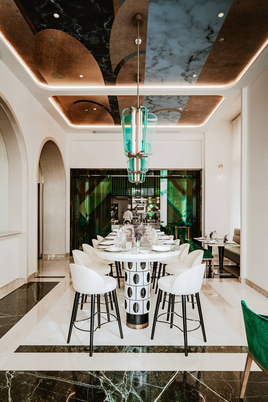 Carlton Cannes, Cannes, France — Modern dining room with turquoise glass pendant, marble ceiling coffers, white barrel chairs, and green-tinted glass kitchen view.