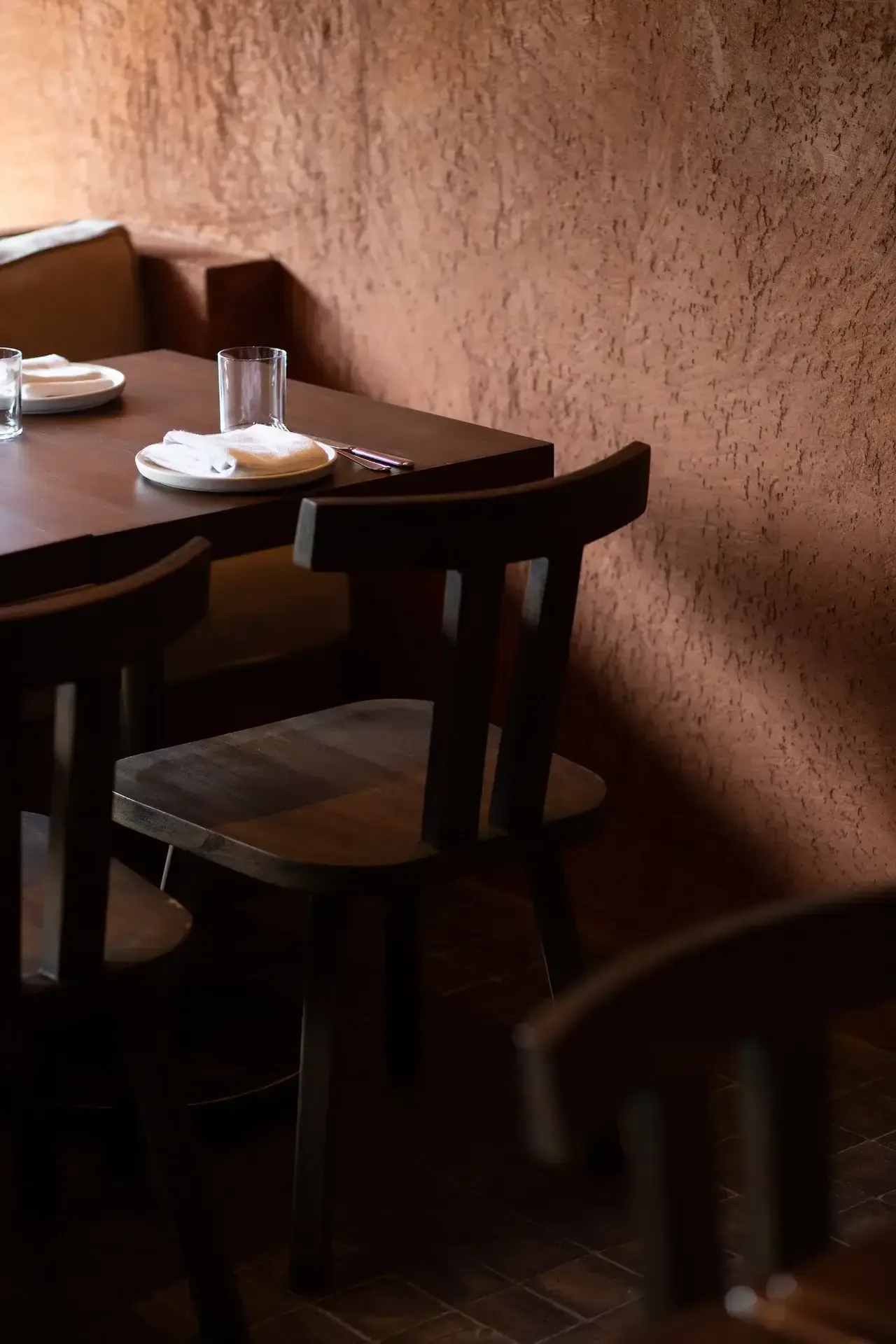 Bar Vera, Asia-Pacific — Intimate dining nook with dark wood table and chairs, textured terracotta walls, minimalist table setting with glass and plate, warm natural light casting shadows.