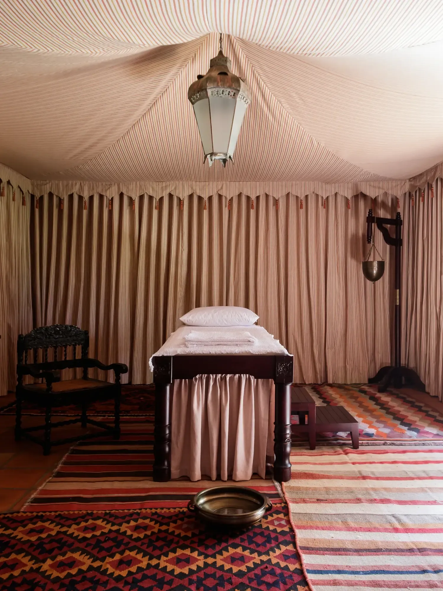 Sterrekopje, Africa — Striped canvas tent interior with dark wood bed, brass pendant lamp, geometric rugs, and draped fabric walls creating intimate safari-style bedroom.