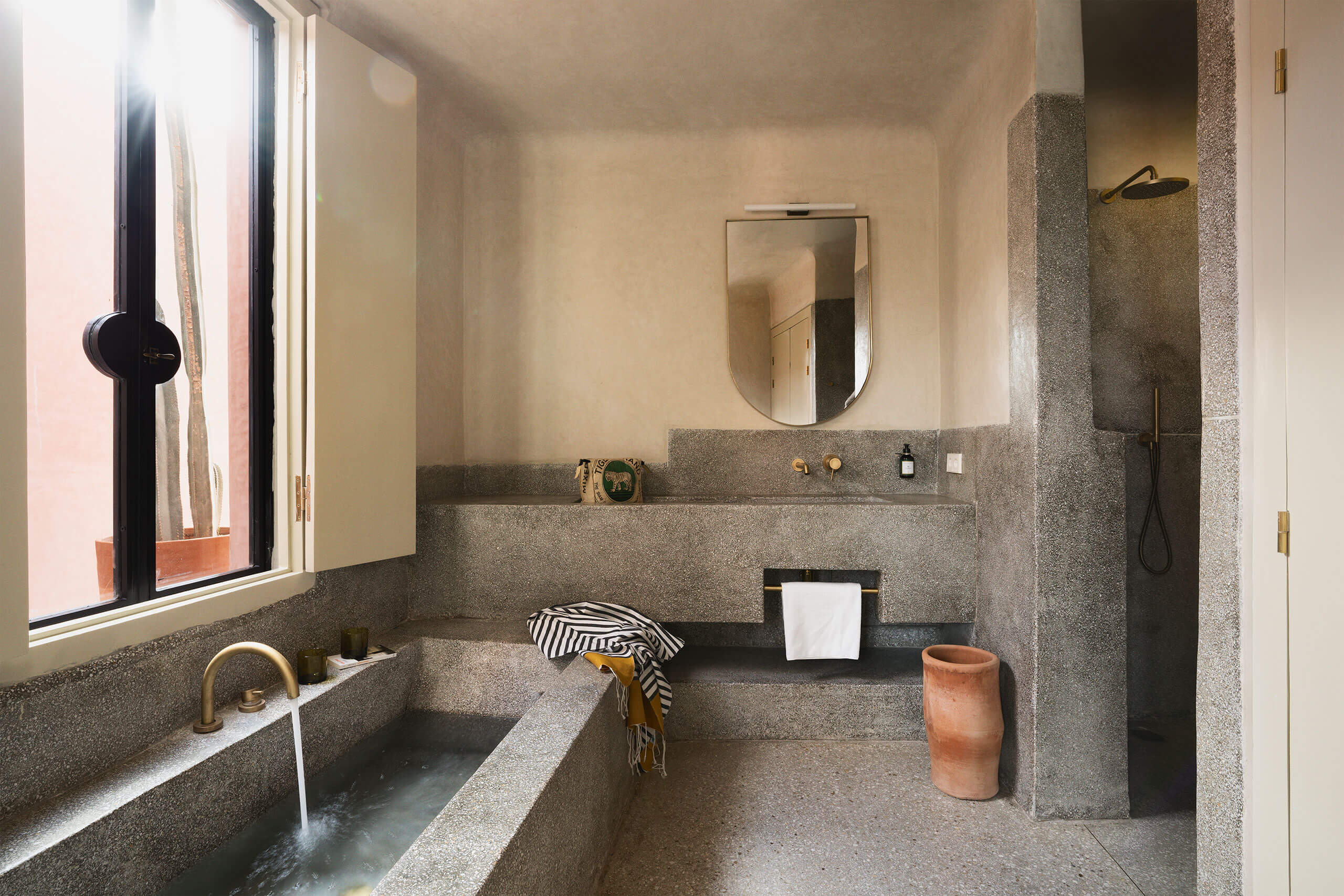 Maison Brummell Majorelle, Africa — Concrete bathroom with terrazzo countertops, sunken soaking tub, minimalist fixtures, and natural light from corner window.