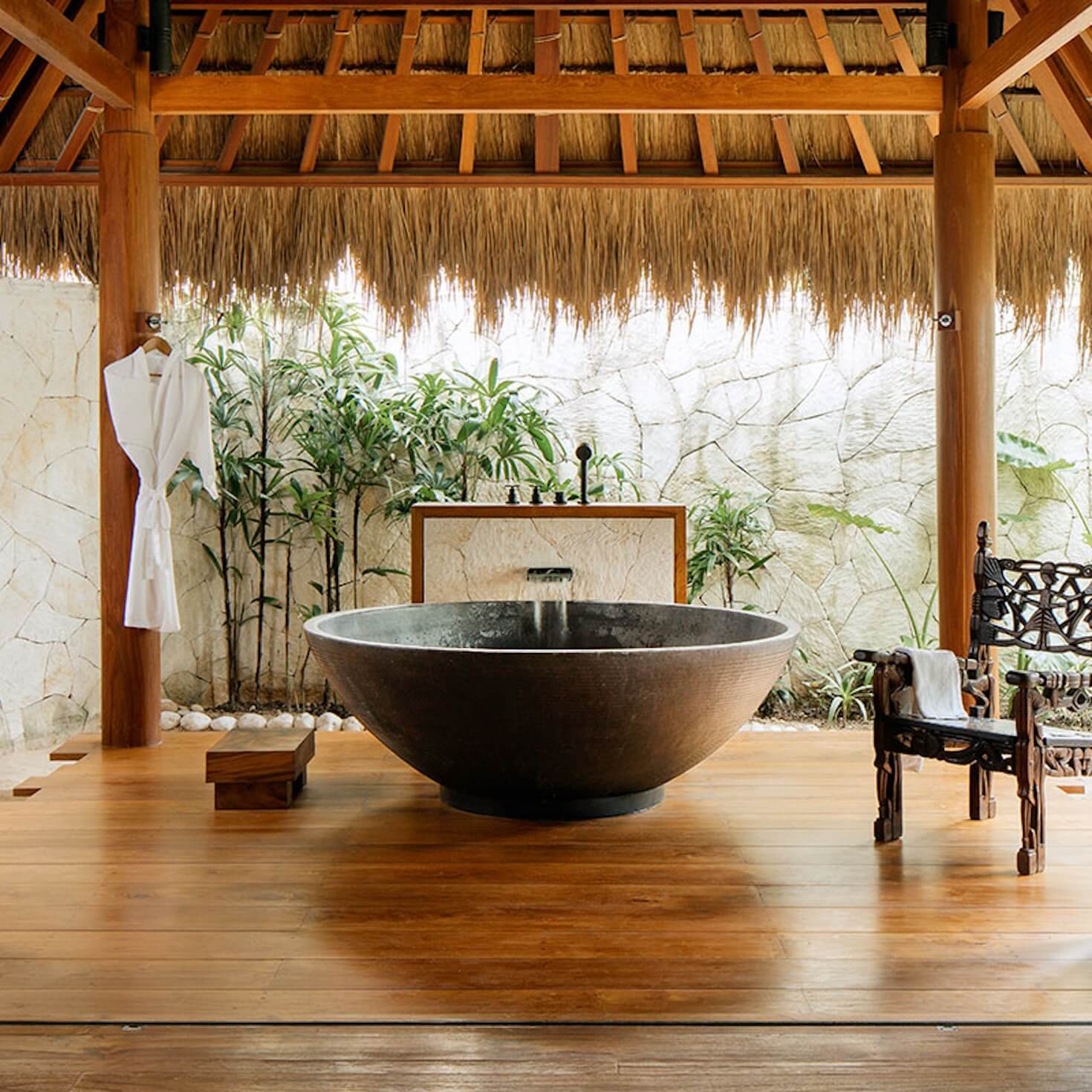 NIHI Sumba, Asia — Sunken stone soaking tub on wooden deck beneath thatched pavilion with bamboo garden and natural stone courtyard wall.
