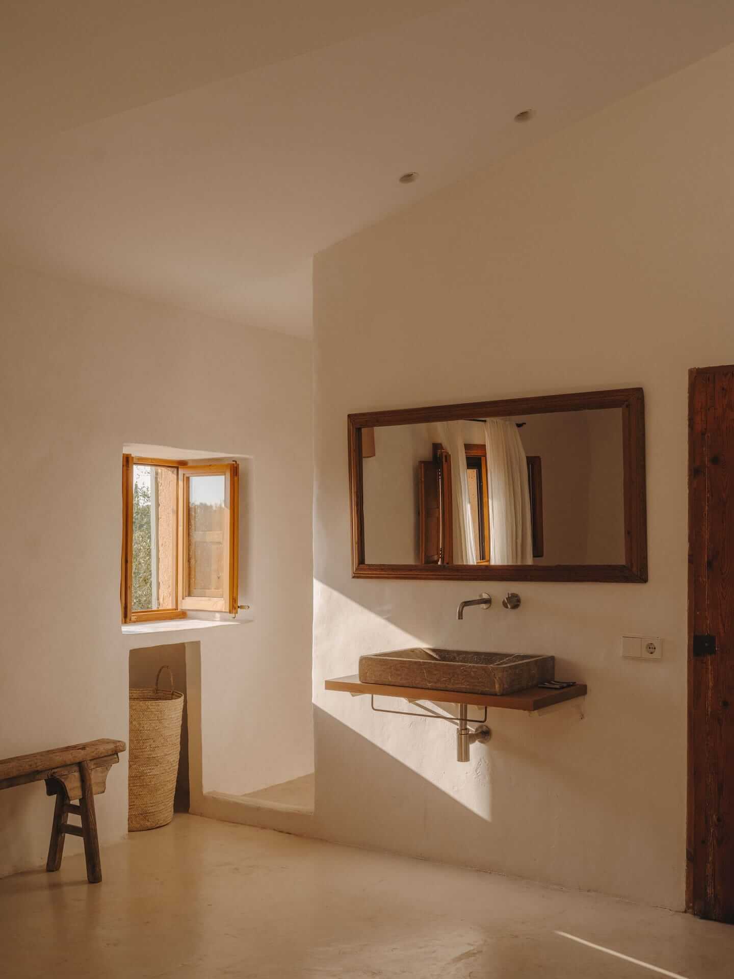 Can Lluïssó, Balearic Islands, Spain — Minimalist bathroom with stone vessel sink, wooden mirror frame, natural light from small window, terracotta tile flooring, whitewashed walls.
