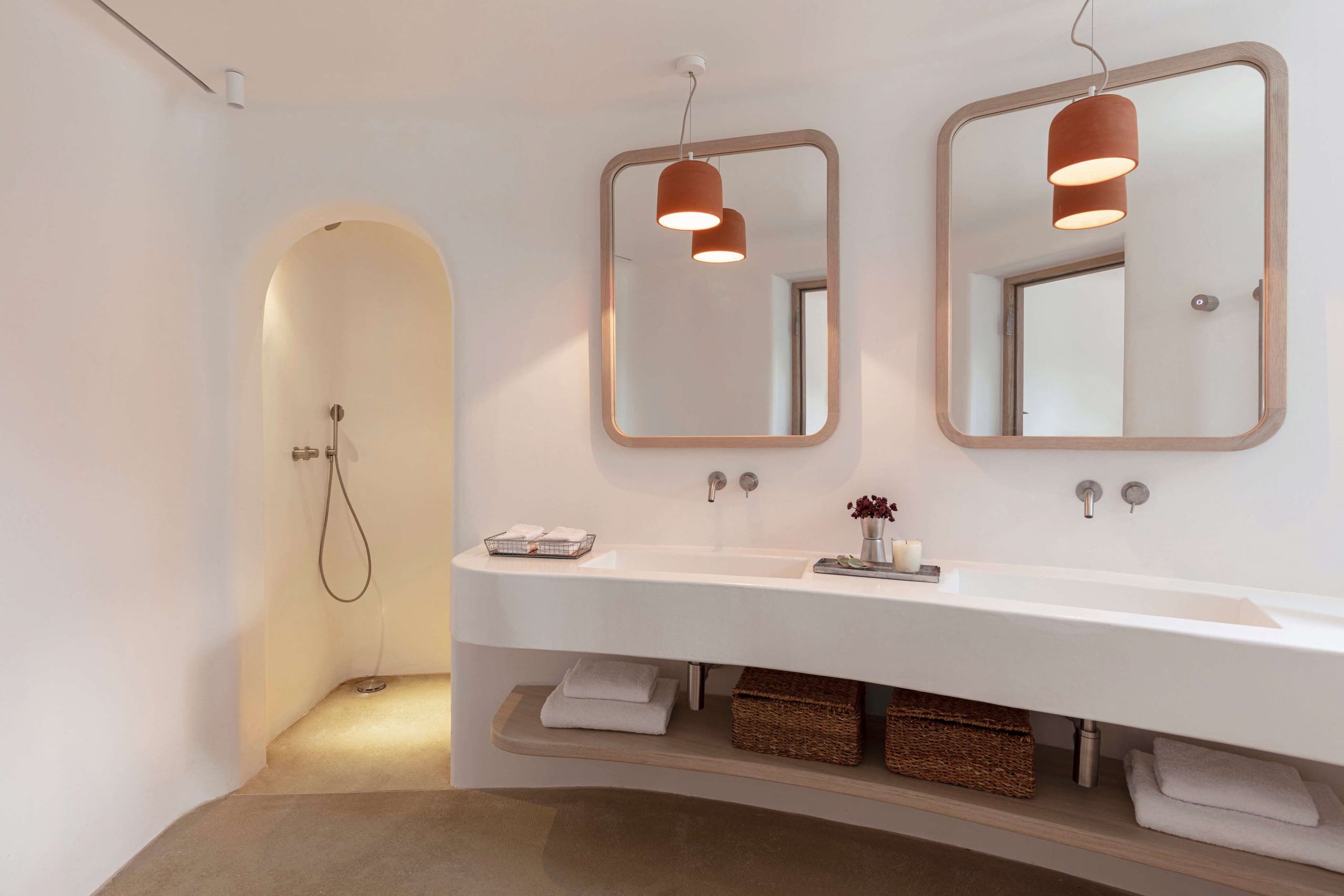 Villa Tama, Europe — Minimalist bathroom with curved archway shower, dual vanity, wood-framed mirrors, terracotta pendant lights, and warm concrete flooring.