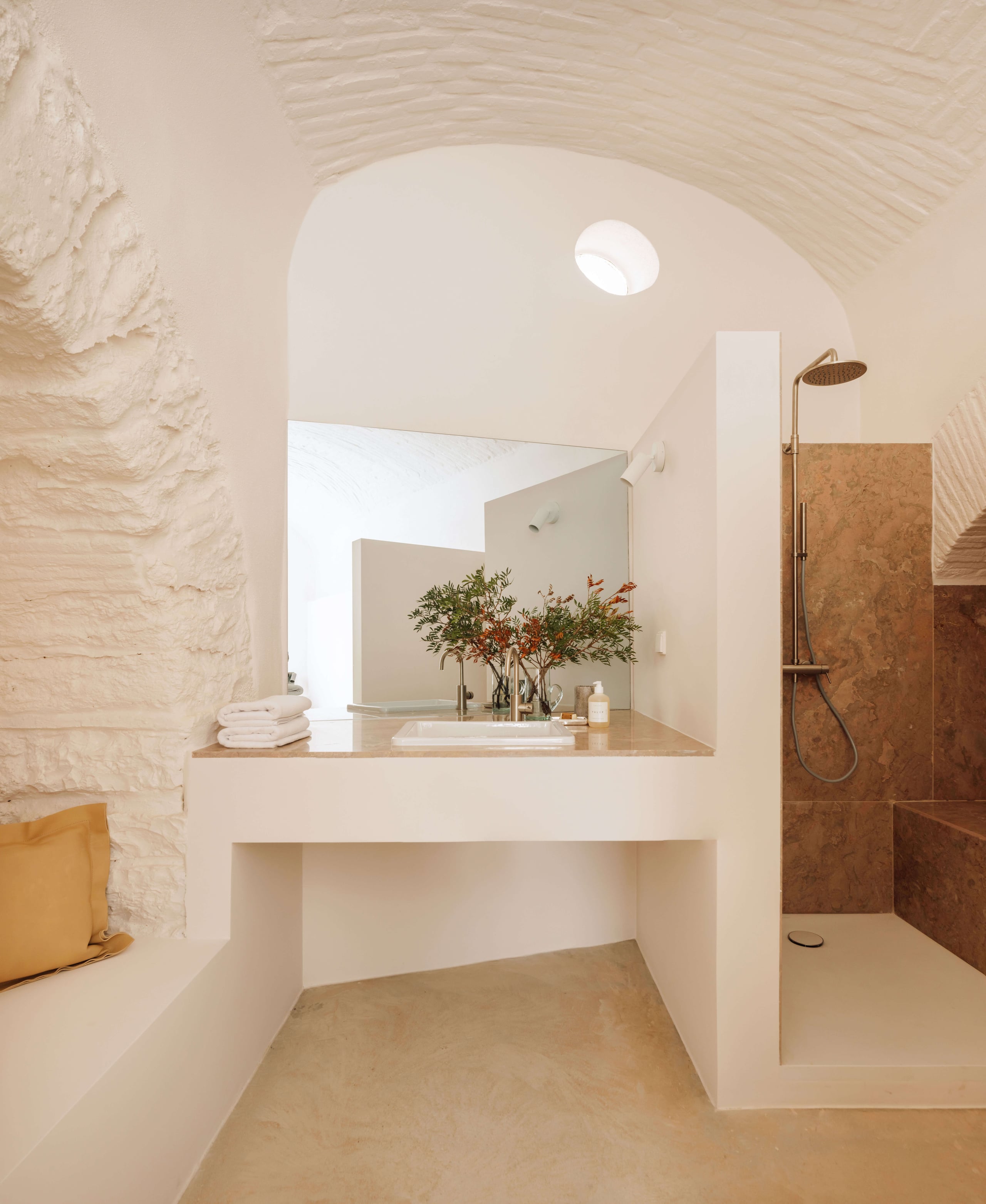 Casa Quatro, Europe — Minimalist bathroom with curved archway, terrazzo vanity, brass fixtures, and rust-toned stone shower featuring natural light and organic flowers.